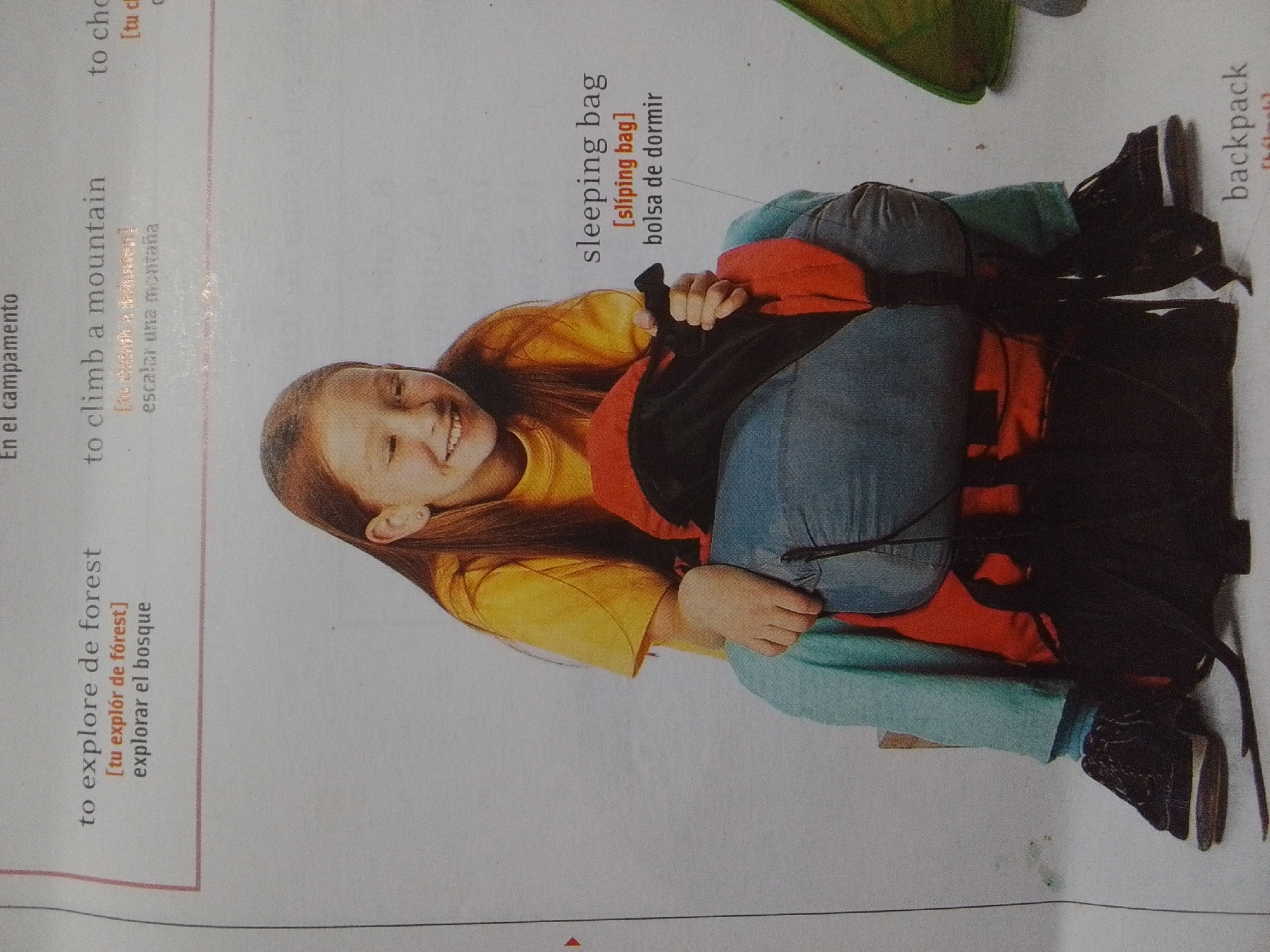 This is a close-up, slightly angled photograph of a young person lying down with a large backpack and sleeping bag. The person is wearing a yellow t-shirt and blue jeans. They have long brown hair and are smiling. The backpack is black and red, and the sleeping bag is red. The background is a white surface, possibly a page from a book or magazine, with text in English and Spanish. The text refers to camping activities like climbing mountains, exploring forests, and mentions a sleeping bag and backpack. The image does not contain any information or visual elements that would allow for identification of the location as La Tagua, Venezuela.