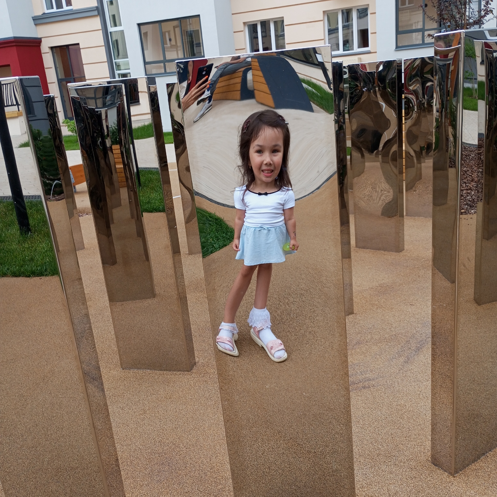 In a city park in Pervomaysky, Russia, a young girl stands amidst a series of polished, vertical metal panels that reflect her image and the surrounding environment. She is wearing a white t-shirt with a black bow at the collar, a light blue skirt, white socks with lace trim, and pink sandals. She is holding a small yellow object in her right hand. The panels distort the reflections, creating a surreal and fragmented view of the girl and the park. In the background, a modern building with beige and white facades and large windows is visible. The ground is covered with a textured tan material, and patches of green grass are present.