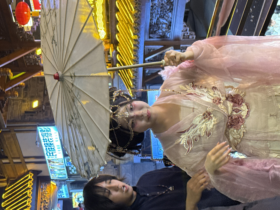 The image captures a vibrant night scene, likely in a street or market setting, featuring two individuals against a backdrop of traditional Chinese architecture and glowing lights. The photograph is oriented vertically, showing the main subject, a woman, in the upper half, and another person, a man, in the lower half.
The central figure is a woman dressed in elaborate traditional Chinese attire. Her face, seen in a slight upward glance towards the viewer, has fair skin, subtle pink blush on her cheeks, and pink lips. Her dark hair is styled intricately with numerous gold-colored ornaments, including a prominent headpiece with dangling chains and beads that frame her forehead and temples. She wears a flowing, light pink or peach-colored dress, possibly a hanfu, with sheer, puffy sleeves. The bodice of her garment is richly embroidered with intricate patterns in gold, white, and maroon or dark pink, resembling floral and feather motifs. She holds a large, light-colored (appears white or cream) paper umbrella with a visible red cap at its center. The umbrella's surface features delicate dark designs, possibly depicting bamboo or other traditional elements, and is held by its handle in her right hand.
Below and slightly to the left of the woman is a man with dark, medium-length hair that frames his face. He is wearing a dark, possibly black, t-shirt. He is looking upwards, towards the woman, with his mouth slightly open, suggesting he might be speaking or reacting to something. His left hand is visible, resting near his side.
The background is a dynamic array of artificial lights and architectural details. The scene appears to be at night, illuminated by numerous bright yellow neon strip lights that outline the edges of buildings or structures, creating a warm glow. Red traditional Chinese lanterns hang in the upper left corner. Various signs with Chinese characters are visible throughout the mid-ground. One prominent sign is illuminated in blue light, displaying several Chinese characters, though they are not fully legible. Other signs are also lit with white or blue light. The architecture in the background features dark wooden elements and intricately carved details, hinting at traditional Chinese building styles.
The overall impression is one of cultural vibrancy and nighttime activity, possibly during a festival or a performance, where traditional aesthetics are highlighted.
