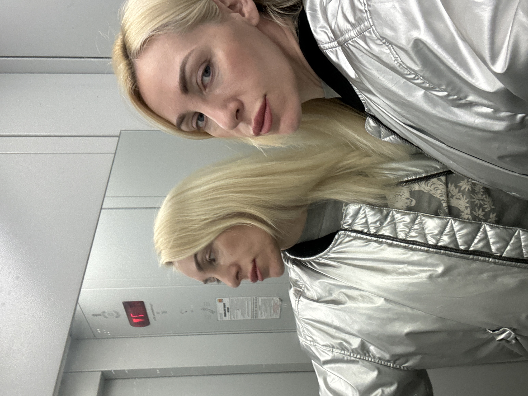 A woman with blond hair is standing in what appears to be an elevator, based on the mirrored view reflecting the elevator panel. She is wearing a silver, metallic-looking jacket with black trim and has light skin. The elevator's digital display reads "17" in red, presumably indicating the 17th floor. There are also control buttons and an informational panel visible on the elevator wall.