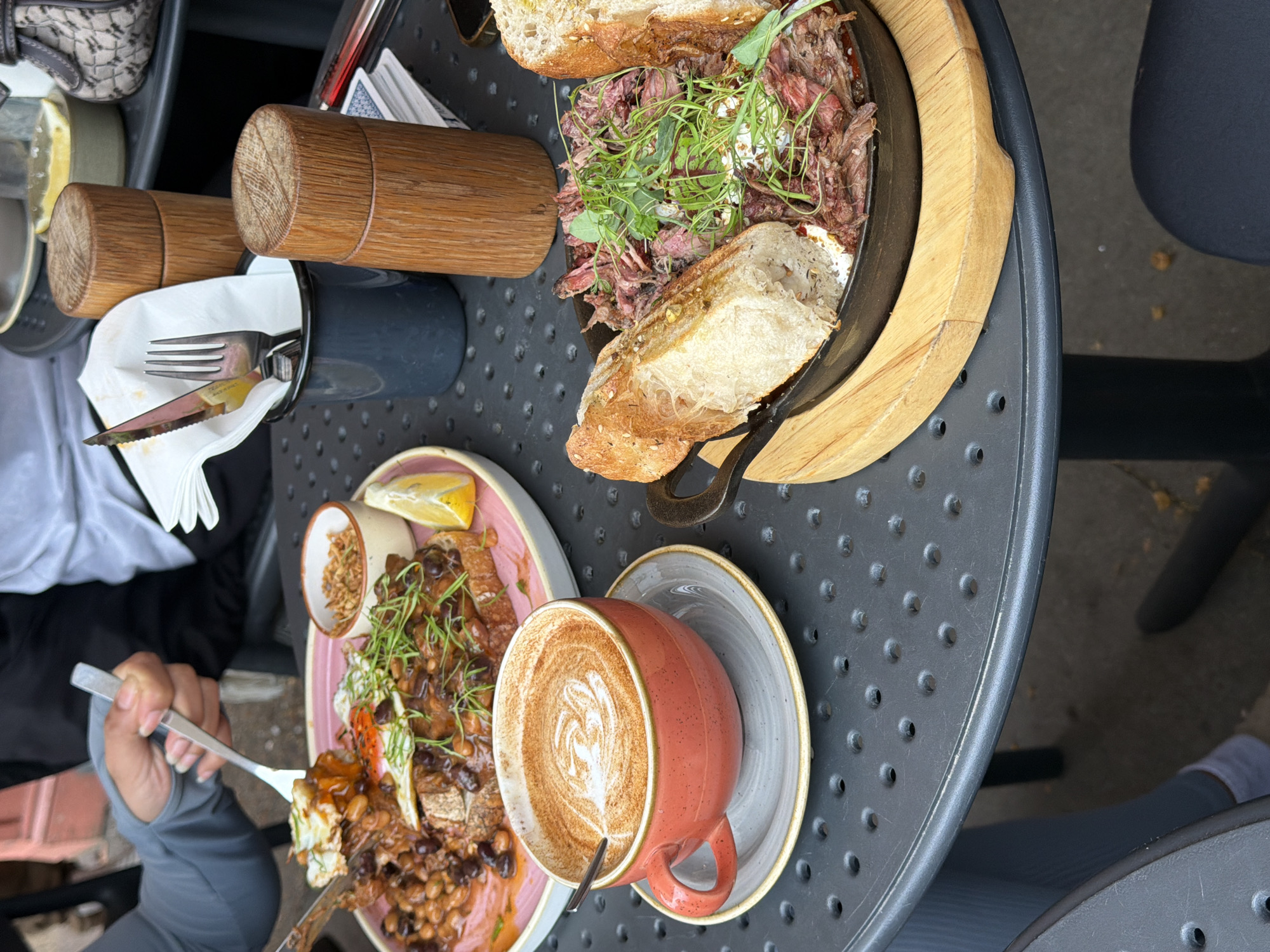 The image captures a table setting with an assortment of food and beverages. There is a cast iron pan with shredded meat, topped with greens and flanked by slices of bread. Beside it is a plate with beans, eggs, fried onions, and greens, along with a small dish of what appears to be a lemon wedge and fried onions. A latte in a pink mug sits on a saucer. Silverware and salt shakers in a dark container are also on the table. The table is dark grey with small perforations. A person is visible with a fork in hand, appearing to be in the process of eating. The location is likely a restaurant in London, United Kingdom.