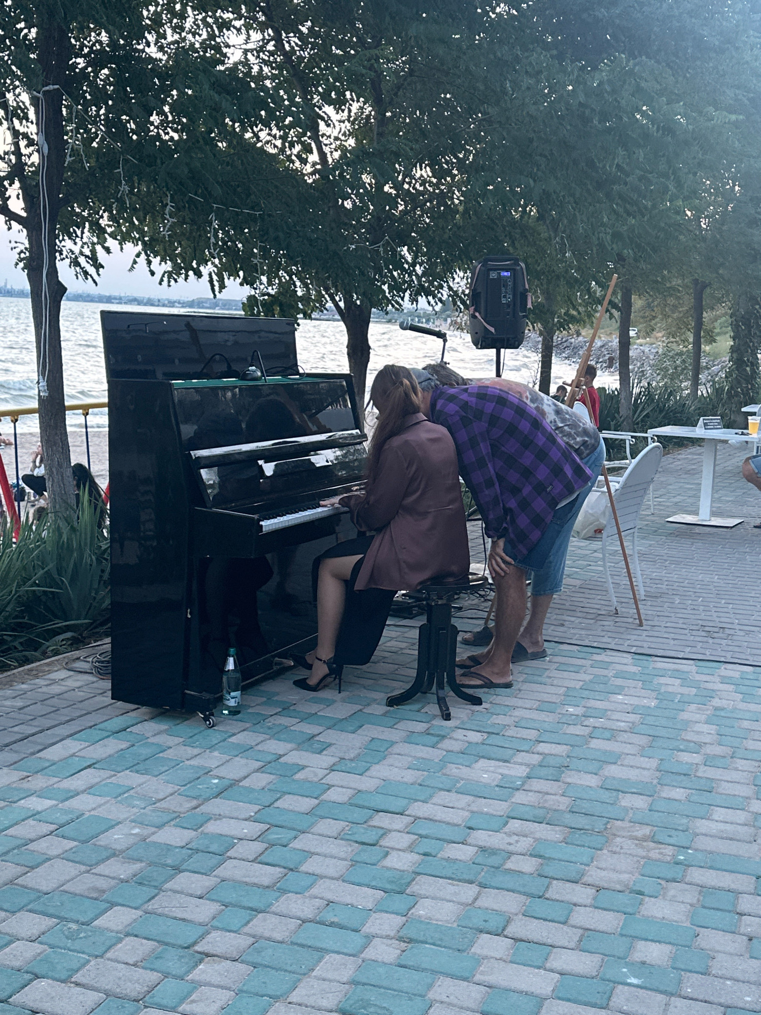 In Kryzhanivka, Ukraine, an outdoor scene unfolds near the sea, featuring a vertical piano set up on a patterned brick patio. A woman with long brown hair, wearing a brown jacket and black high heels, sits on a stool and plays the piano. A man, clad in a purple plaid shirt and denim shorts, leans over the piano, appearing to interact with the woman or the instrument. Behind them, a portable speaker is positioned on a stand, and further in the background, trees line the waterfront, with a glimpse of the sea and a distant shoreline. A white chair and a table with a glass are visible to the right, suggesting a public or dining area. The atmosphere appears relaxed, with the piano performance taking place in a scenic outdoor setting.