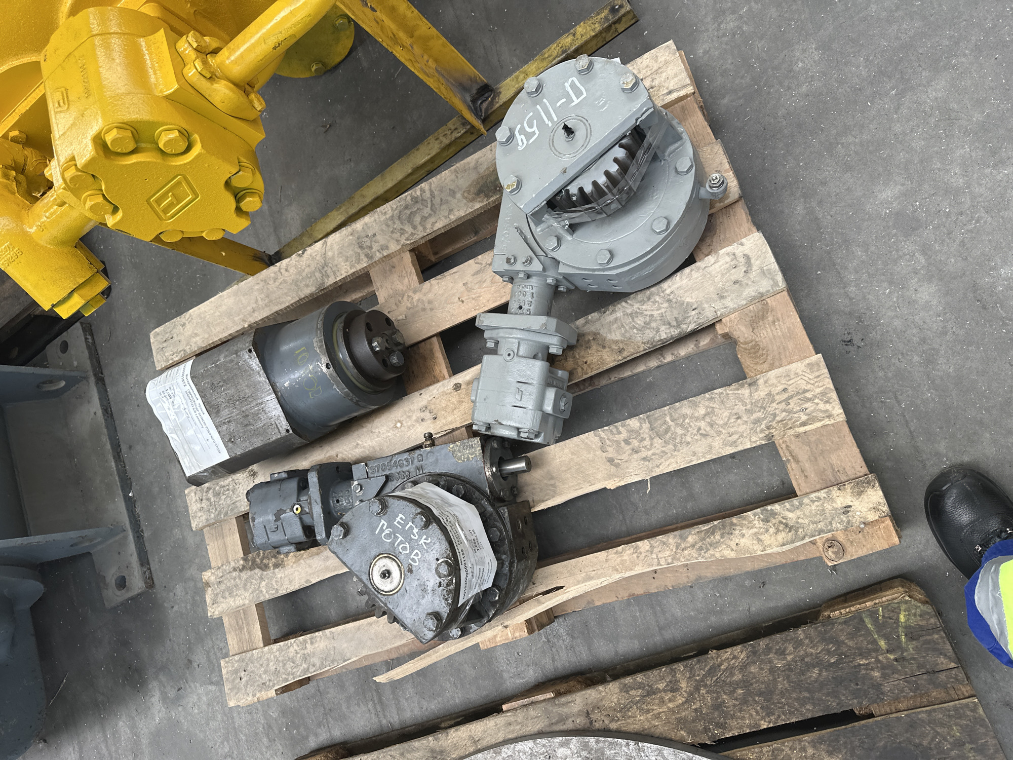 An overhead, slightly angled medium shot shows industrial equipment on wooden pallets on a concrete floor. In the foreground, on a pallet, are two gray hydraulic components. The one on the left is a gearbox or pump with a label indicating "10108". The one on the right is a different hydraulic motor, possibly a pump, with a label visible that says "DT-11-1159". To the right of these, a large yellow industrial piece of machinery is visible, with a central yellow shaft and two large yellow circular housings. The background shows another piece of gray industrial equipment on a pallet. A person's lower leg and foot, wearing a black work boot and blue and yellow safety pants, are visible in the bottom left corner of the image, standing near the wooden pallets. The environment appears to be a warehouse or industrial storage area, with a concrete floor and some industrial equipment visible in the background. No specific location details about the city of Hora, Ukraine are discernible from the image.