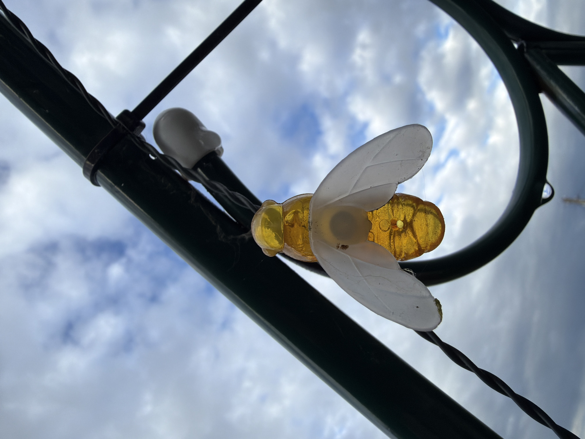 A low-angle shot of a translucent yellow and white bee ornament affixed to a dark green metal structure with dark wires. The ornament is facing the viewer with its wings spread. The background features a sky filled with streaky clouds. The location is Unknown, Unknown.