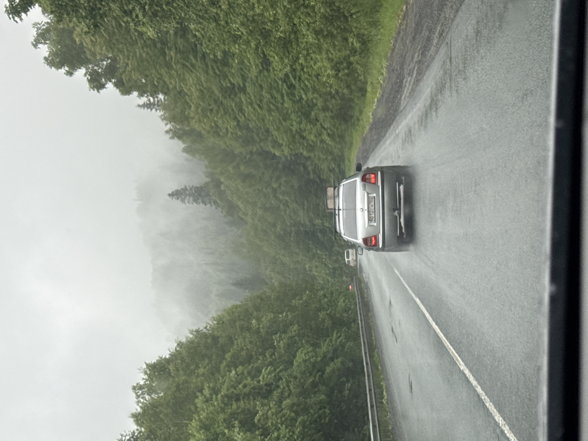 The image depicts a road scene in Brishbash, Russia, characterized by overcast skies and a light rain. A silver sedan is driving away from the camera on a wet, two-lane road bordered by dense green forest. The road surface appears to have some potholes and is covered in a thin layer of water. The forest on either side of the road is lush and extends up a misty, forested hillside. In the distance, other vehicles can be seen on the road. The overall atmosphere is subdued and damp due to the weather.
