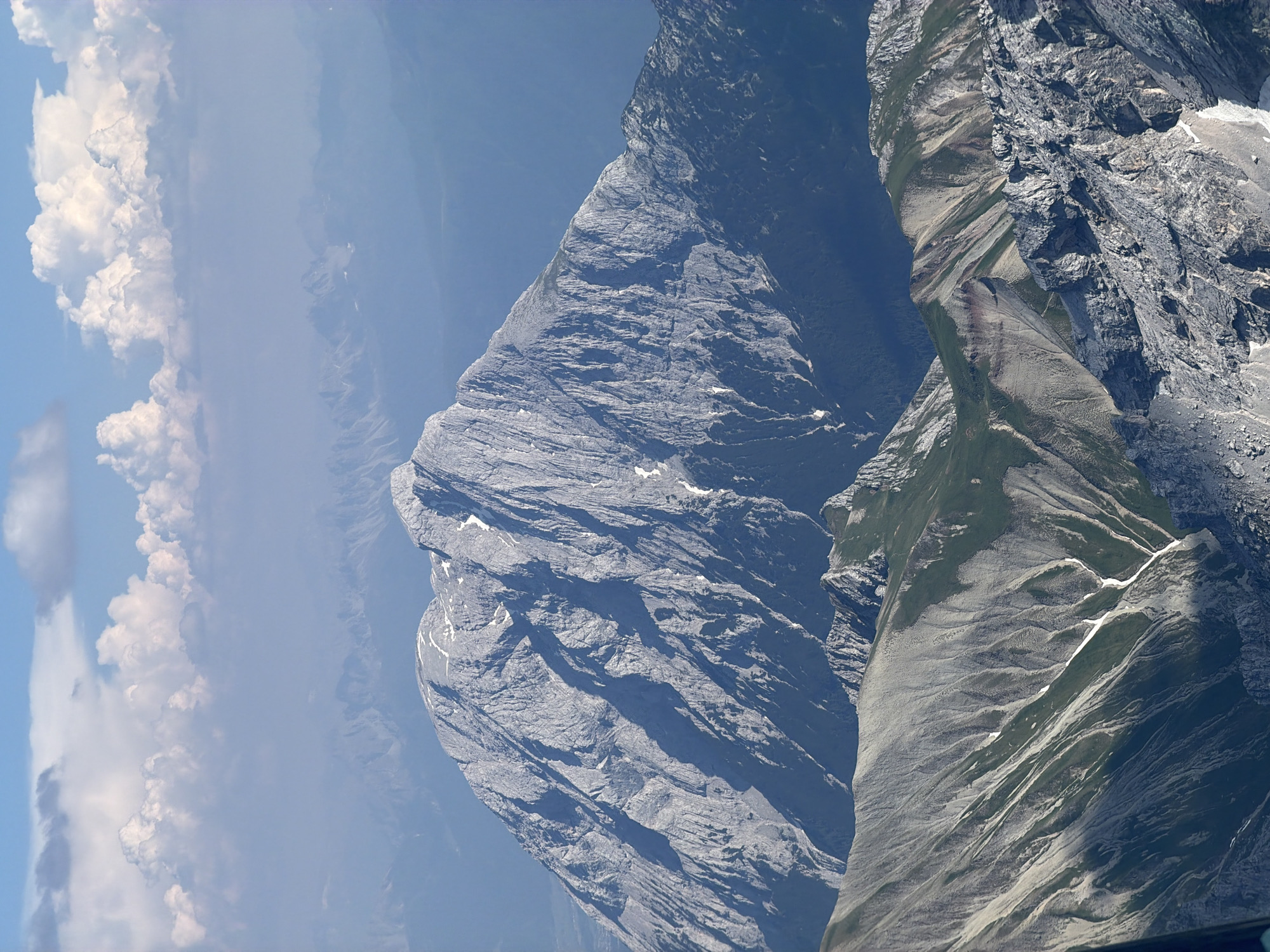 This media file shows a dramatic, high-angle aerial view of a rugged mountain landscape near Garmisch-Partenkirchen, Germany. The dominant subjects are massive, imposing rock formations and steep mountain slopes, characterized by deep ravines, sharp ridges, and weathered surfaces. Patches of snow are visible on some of the higher elevations, indicating a cold or alpine environment. The lower slopes are partially covered in green vegetation, suggesting a transition from alpine to subalpine zones.
The scene is bathed in bright sunlight, casting sharp shadows that emphasize the contours and texture of the mountains. The sky is a clear, deep blue in some parts, but also features scattered white clouds, particularly towards the left side of the frame. These clouds add depth and suggest varying atmospheric conditions. The overall impression is one of vastness and natural grandeur, with the immense scale of the mountains dwarfed by the expansive sky.
There are no people or man-made objects explicitly visible in the frame. The activity or event depicted is simply the observation of the natural mountain scenery from a high vantage point, likely from an aircraft or a very high peak. The time of day appears to be midday or early afternoon, given the strong sunlight and pronounced shadows. The location context of Garmisch-Partenkirchen places this scene within the Bavarian Alps, known for their dramatic topography. No visible text is present in the media file.