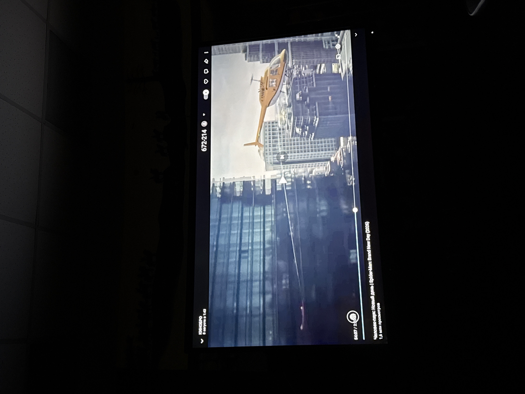 The image shows a television screen displaying a video player interface with a scene from a movie. In the video, a yellow helicopter flies past modern skyscrapers. The video player shows a timestamp "04:07 / 2:30:27" and the title "Человек-паук: Новый день | Spider-Man: Brand New Day (2026)" with "1,6 млн просмотров" which translates to "1.6 million views". The top left corner of the screen shows "КИНОБРО" and "8 августа в 1:43". The scene is viewed in a dark room, likely a home theater or a dimly lit room, with the ceiling visible in the upper portion of the image. The location in the city of Nyurba, Russia, is not directly discernible from the image content, as it depicts a city scene that could be in many urban locations.