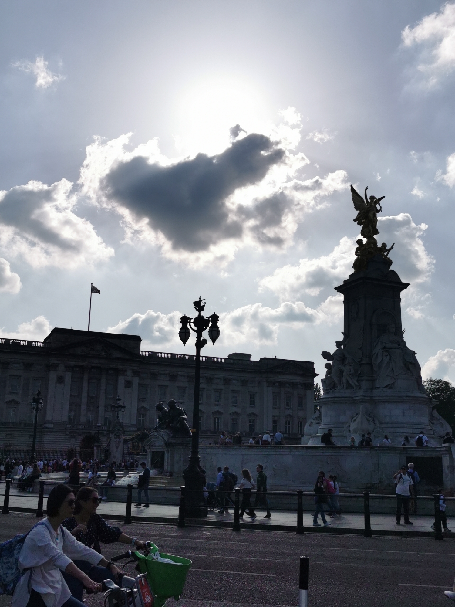 This daytime photograph captures a bustling scene in front of Buckingham Palace and the Victoria Memorial in London, United Kingdom. The grand, neoclassical architecture of Buckingham Palace stands in the background, appearing silhouetted and dark against the bright sky. A flag is flying from the palace roof.
Prominently featured on the right side of the image is the Victoria Memorial, a large, multi-component monument. Its base consists of white marble sculptures, while a golden winged figure, likely Victory, crowns the top, catching the sunlight.
Numerous people populate the scene. Many pedestrians are visible on the pathways surrounding the memorial and in the distance near the palace, suggesting a busy tourist location. In the immediate foreground, two women are cycling on a road. One woman wears a white shirt and sunglasses, while the other, dressed in a darker patterned top and sunglasses, rides alongside, her bicycle equipped with a bright green basket. Ornate black lampposts and railings line the road, separating it from the pedestrian areas.
The sky is dramatic, filled with large, bright clouds. The sun, partially obscured by these clouds, creates a strong backlight effect, casting many elements, including the palace, the memorial, and the people, into silhouette. This backlighting results in a high-contrast image, emphasizing the bright sky and the shapes of the subjects. The weather appears to be partly cloudy but bright. No discernible text is visible.