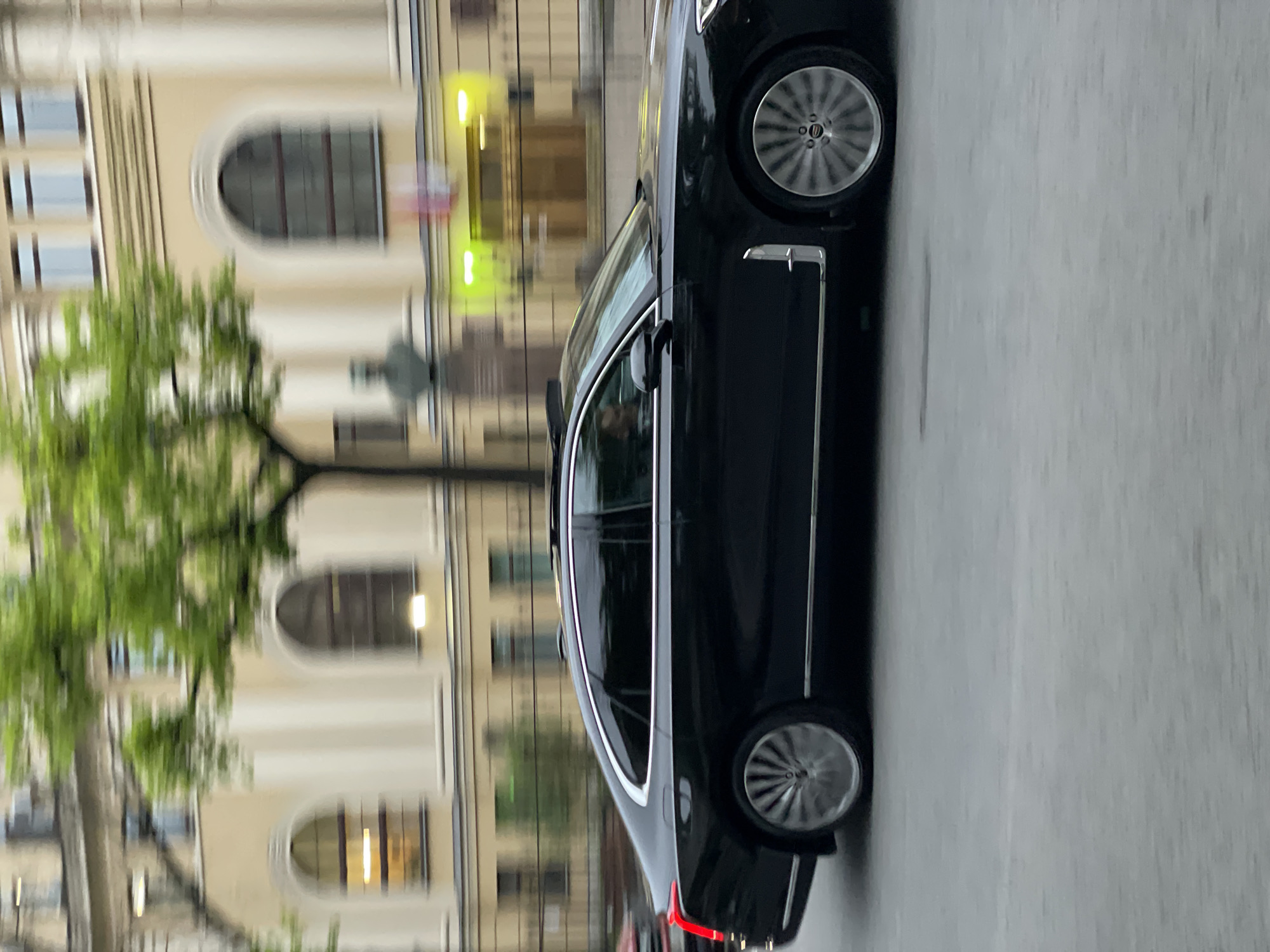 A black car is parked on a street. The building in the background suggests this location may be in Saint Petersburg, Russia. The car is parked on the right side of the road. The building behind it has arched windows and is of light-colored stone. A tree with green leaves partially obscures the building. The street appears to be wet, possibly from rain.