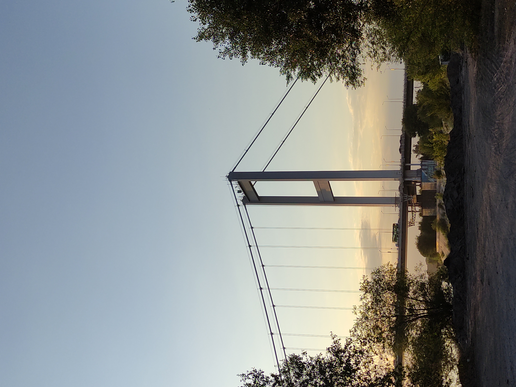 The image depicts a suspension bridge spanning a body of water, likely a river. The sky is clear and pale blue, with a gradient of yellow and orange hues near the horizon, indicating either sunrise or sunset. In the foreground, a rough, dirt surface is visible, with piles of dark soil. To the left and right sides of the frame, dense green foliage from trees partially obscures the view.
The suspension bridge features a prominent, tall, grey concrete tower. Two thick, dark cables run from the top of the tower, converging with similar cables from an unseen tower to support the bridge deck. The bridge deck itself is visible in the mid-ground, with vehicles, including a green bus, traveling across it. Streetlights are also present along the bridge. Further in the background, another bridge or elevated structure is visible, also with lights.
The scene appears to be located in the city of Semey, Kazakhstan, as the bridge is identifiable as the Old Semey Bridge over the Irtysh River. The overall atmosphere is serene, with the natural light casting a warm glow on the scene. There are no clearly visible people in the image, apart from a few indistinct figures standing at the top of the bridge tower, who appear to be construction workers or maintenance personnel.