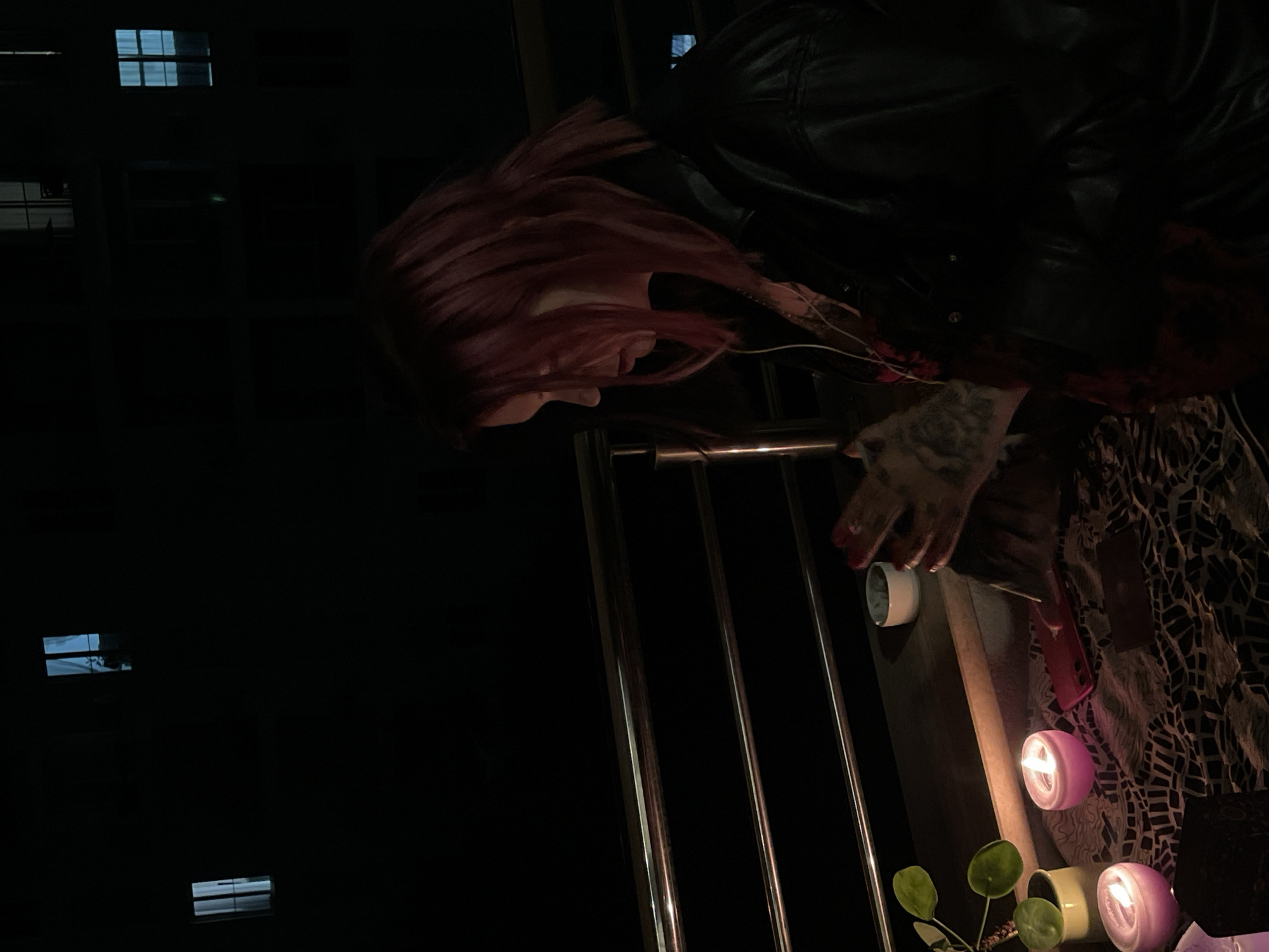 The image displays a nighttime scene on an outdoor balcony in Boreti, Montenegro. A person with pinkish-red, shoulder-length hair is visible in profile, facing left and looking downwards. They wear dark outerwear over a patterned garment, with white earphones around their neck. Tattoos are present on the person's right forearm and hand, which are positioned above a patterned cloth covering a surface.
On this surface, from left to right, are a potted plant with round green leaves, two lit pink candles in rounded holders, a red-cased smartphone, a dark rectangular object, and a small white bowl. A metal balcony railing with multiple horizontal bars is in the midground. The background consists of dark building silhouettes containing several illuminated windows, some emitting a bluish light. The primary light sources are the candles in the foreground and the distant building windows.