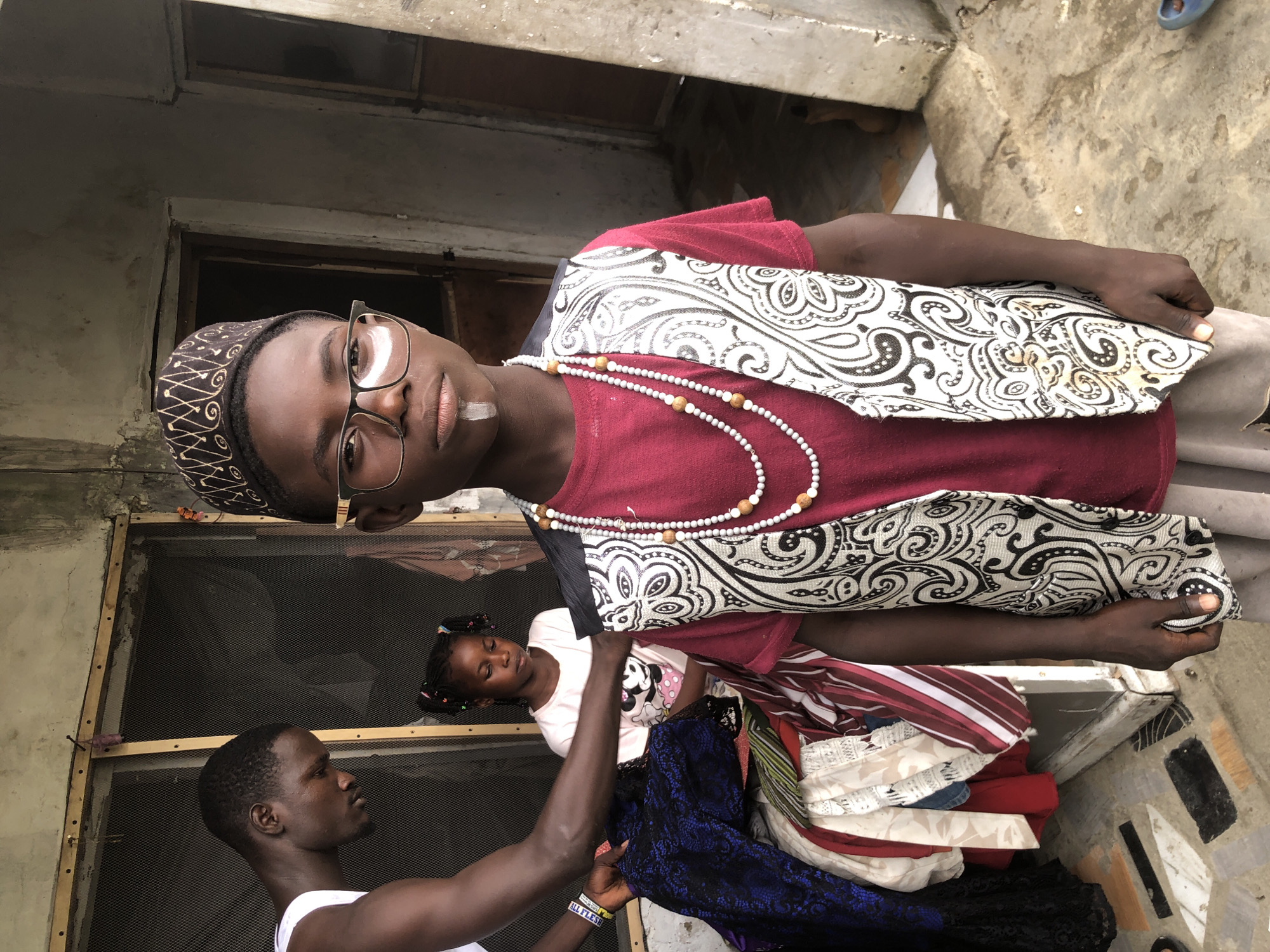 In Ado, Nigeria, a young boy stands facing the camera, dressed in a maroon t-shirt, a white and black patterned vest, and a patterned Kufi hat. He wears glasses with gold rims and has a white stripe painted vertically on his upper lip and chin. A beaded necklace adorns his neck. To his left, a man wearing a white tank top sorts through a pile of colorful clothing. In the background, a young girl with braided hair sits looking down, wearing a white t-shirt with a Mickey Mouse print. The scene appears to be outdoors, possibly in a courtyard, with a rough concrete floor and weathered building structures. The overall impression is a candid moment captured in a domestic or semi-public setting.