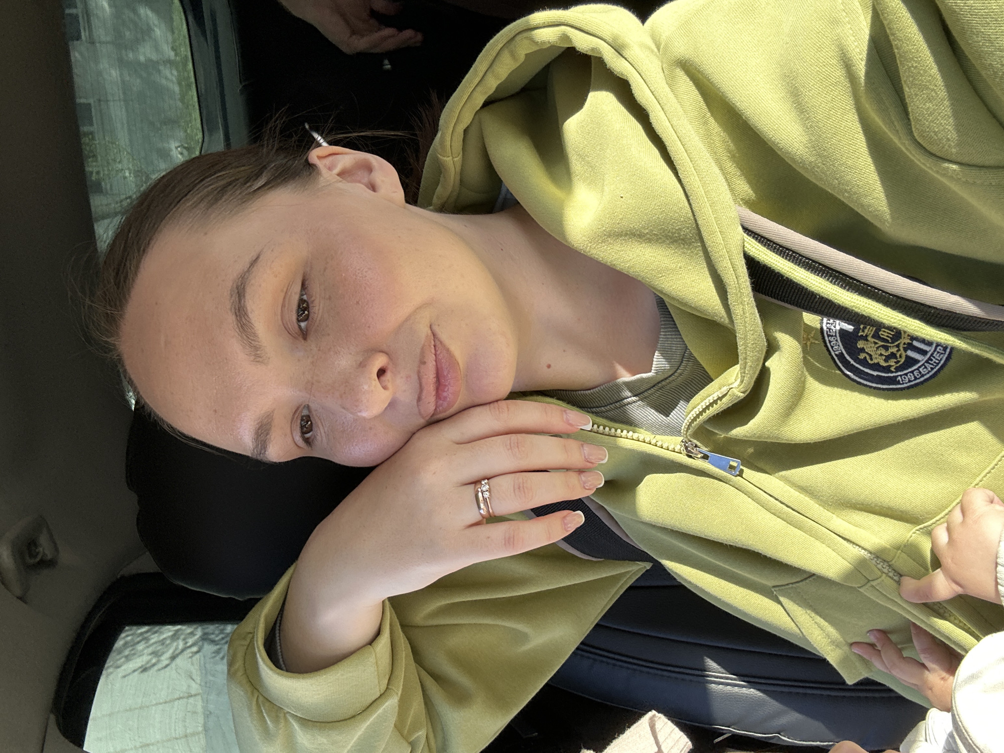 A person lies in a car wearing a green hoodie, likely a passenger as they are reclined against a headrest next to a car window. A child’s hand, presumably in the same car, is on the zipper of the hoodie. The ring on the person's finger is visible. The city of Chaikovsky, Russia is not identifiable within the image.