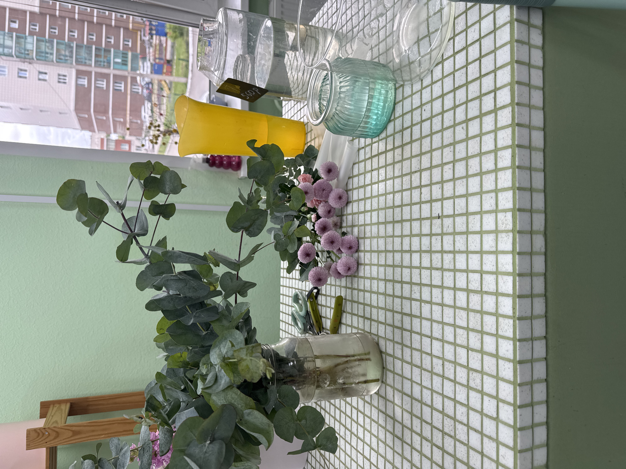 The image depicts a still life on a tiled surface, likely a countertop or windowsill, indoors.
Subjects: A bouquet of eucalyptus branches and small purple flowers in a clear glass jar. Additional purple flowers are arranged nearby. A pair of scissors and a pen-like object lie on the tiled surface. Several glass jars of various shapes and sizes are present: clear cylindrical jars, one with a price tag ("560 P"), and a small turquoise glass jar. A yellow plastic container is also present.
Environment: The background includes a window showing an urban landscape, possibly a residential area, with multi-story buildings. The scene is located in Izhevsk, Russia. The surrounding environment is light green, likely a wall. The tiled surface is white with green grout lines.
Objects: Eucalyptus, purple flowers, clear glass jar, turquoise glass jar, yellow container, clear cylindrical jars, scissors, pen, tiles, and windowsill or countertop.
The image presents a collection of everyday objects arranged in a decorative manner. The inclusion of the building and the text "Izhevsk, Russia" gives context to the location.