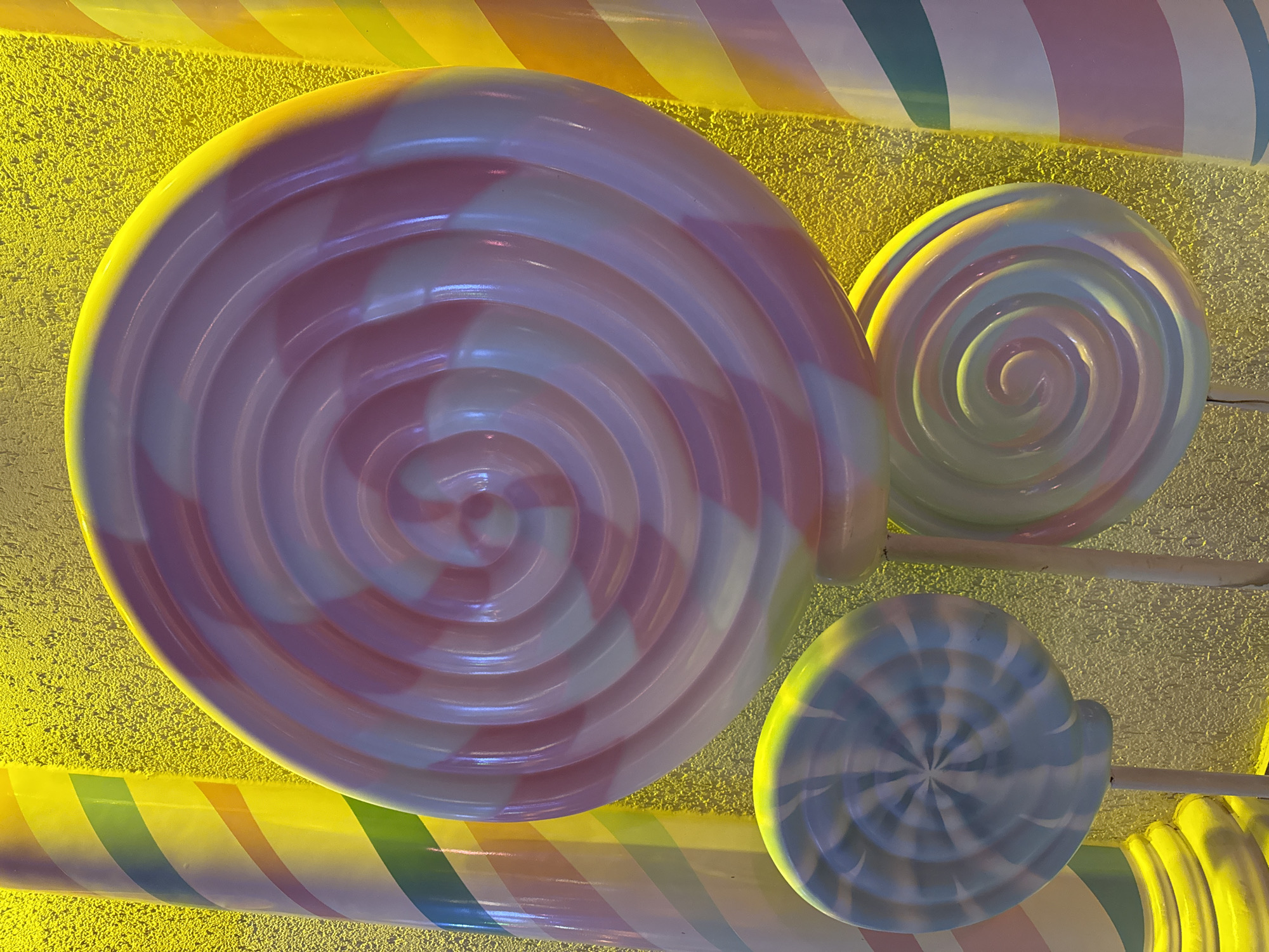 The image displays a close-up of several oversized, decorative lollipops against a textured yellow background. The lollipops are round with swirled patterns in various color combinations, including pink and white, and pale blue and white. Some are also positioned next to large, striped candy cane-like pillars in yellow, orange, green, and white. The lighting is warm and vibrant, creating highlights on the glossy surfaces of the candy decorations. The scene appears to be an outdoor display, possibly in a theme park or a festive area in Bodrum, Turkey, given the bright, cheerful aesthetic often associated with tourist destinations. No people are visible in the image, and the focus is entirely on the candy-themed decorations.