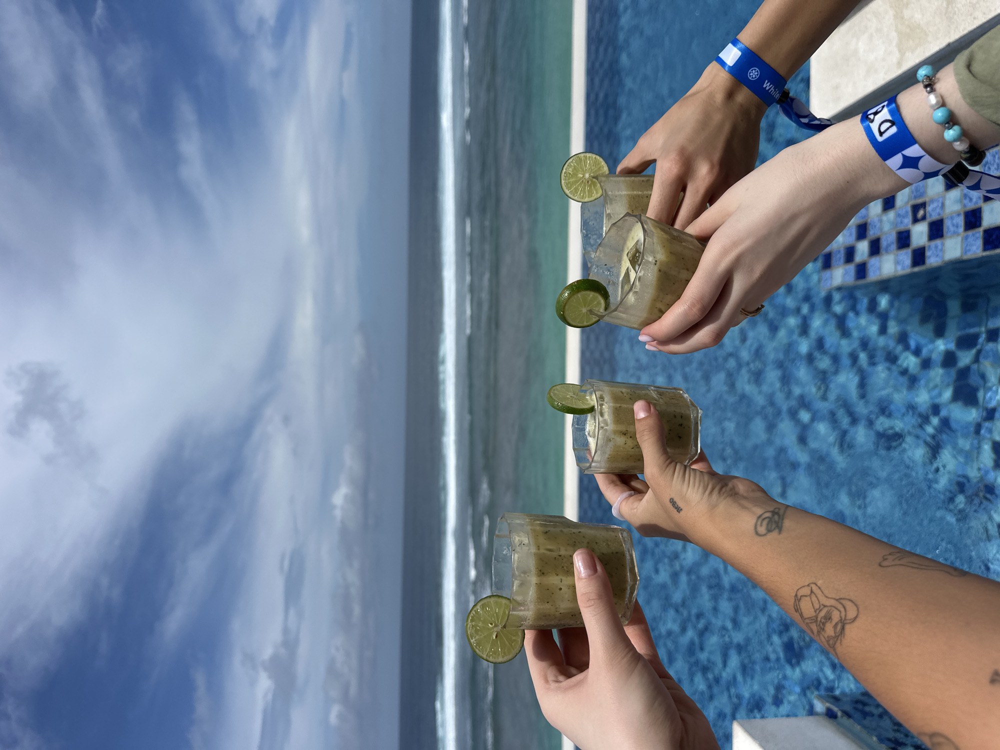 Three individuals hold glasses of beverages with lime garnishes, potentially smoothies or cocktails, against a backdrop of ocean and sky. The location appears to be near or within a poolside area featuring blue and white tiles. Each person's arm and hand are visible. Two of the individuals have blue wristbands, one with the word "Whitley". The wrist with the wristband with the word Whitley also has a bead bracelet with turquoise colored beads. The third person's arm is tattooed. The ocean has visible wave patterns. The sky is blue with white clouds. It is likely the individuals are engaging in a celebratory toast. The image appears to be taken at an elevated location overlooking the ocean in Kuta Selatan, Indonesia.
