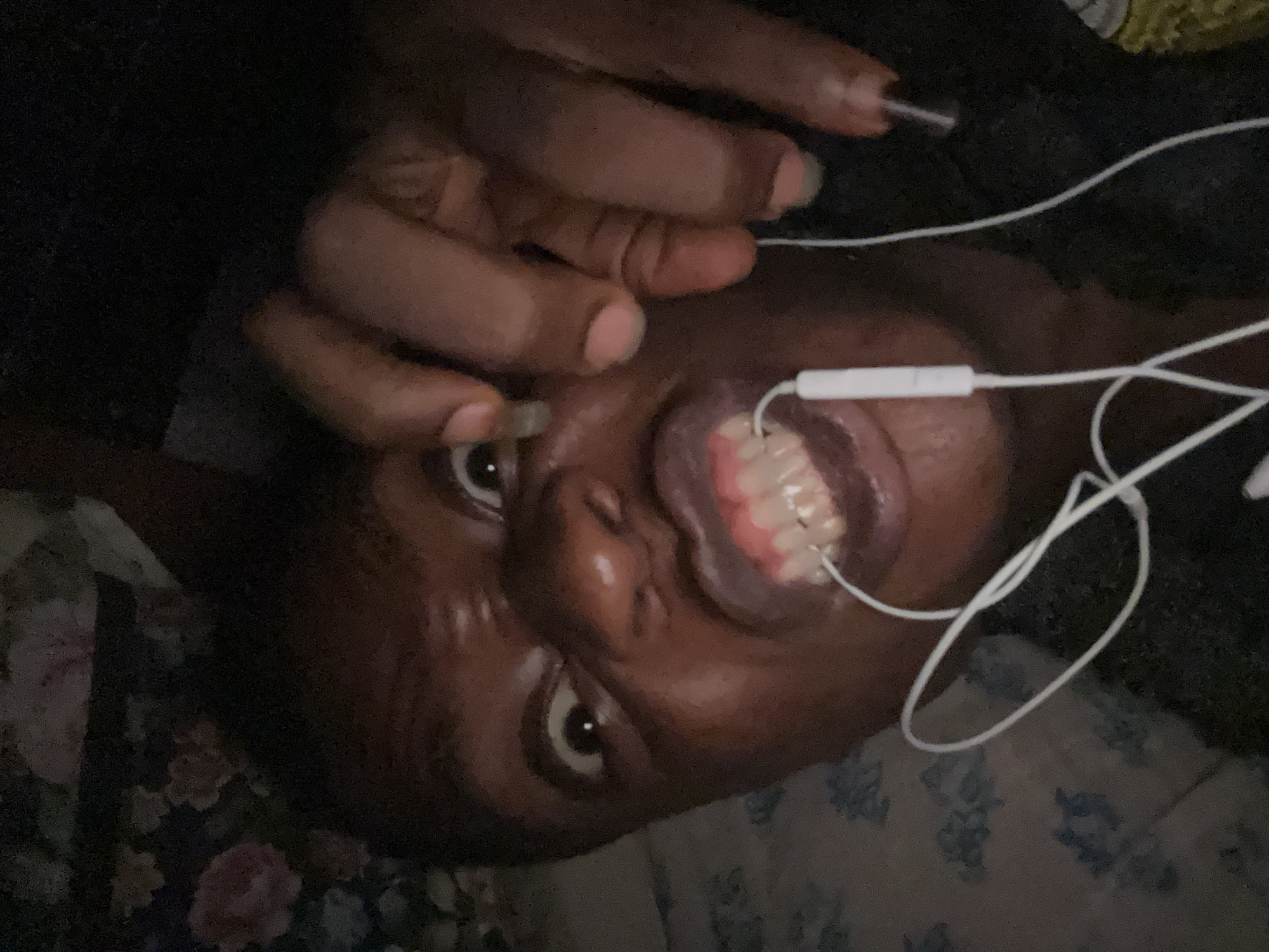 A person with dark skin and short, dark hair is lying on a floral patterned surface. They have white earphones plugged into their mouth. Their hand is raised near their face. The location is unknown.