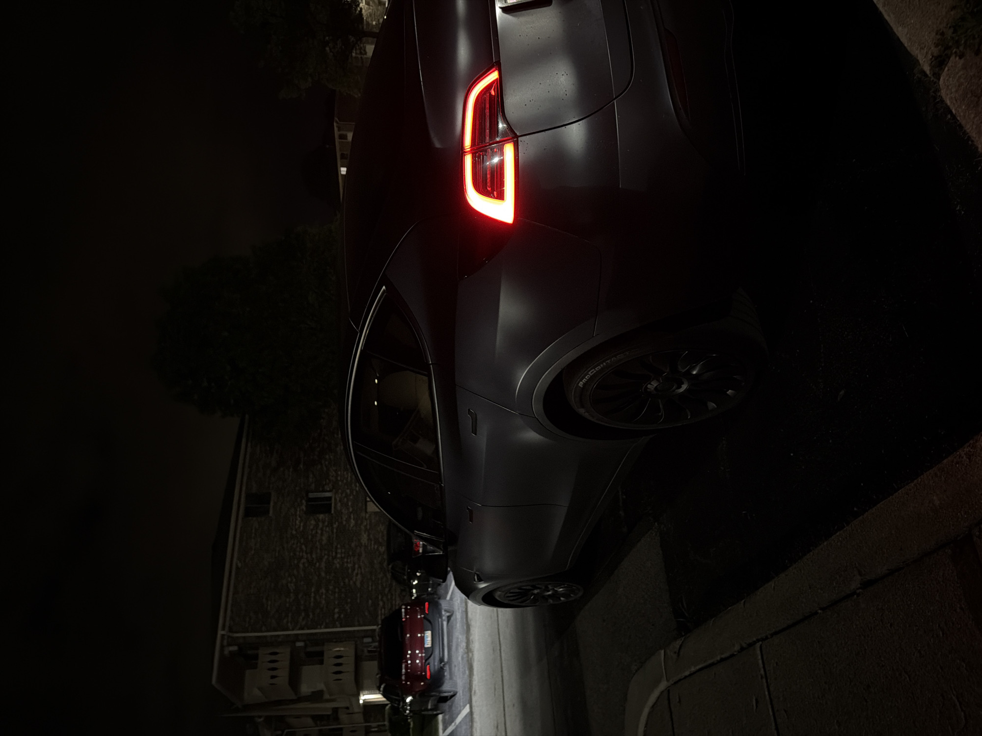 This is a nighttime, eye-level, medium shot of the rear passenger side of a dark gray Tesla Model Y parked on asphalt next to a curb. The car's brake lights are illuminated, emitting a bright red glow. In the background, to the left of the Tesla, a dark red SUV is parked in a lit parking spot. Further in the background, a brick apartment building with balconies and vine-covered walls is visible. Trees are present on the right side of the frame. The overall lighting suggests it is dark, with artificial lights from the apartment building and parking lot illuminating portions of the scene. There are no people visible in the image. The location is identified as Vernon Hills, United States of America.