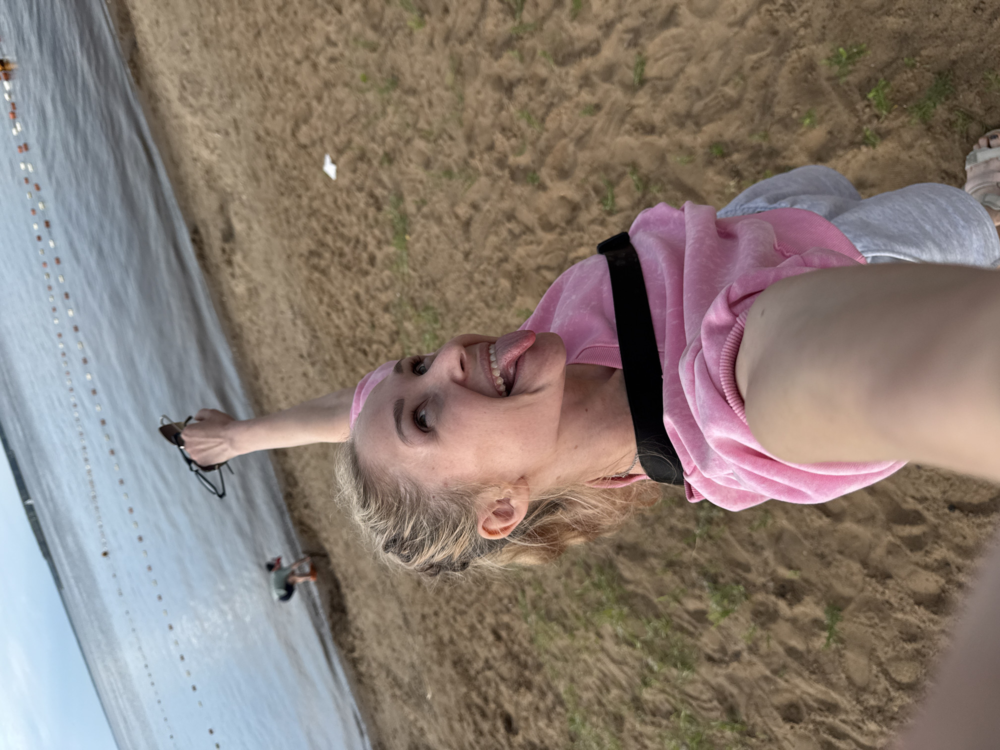A young woman is taking a selfie at a sandy beach on a cloudy day. She is wearing a pink hoodie and light gray shorts. Her tongue is sticking out and she is looking up and to the side. In the background, the water is calm and there is a rope with buoys extending into it. A person is bent over near the water's edge. The location is the Kama River embankment in Chaikovsky, Russia, a popular recreational area.