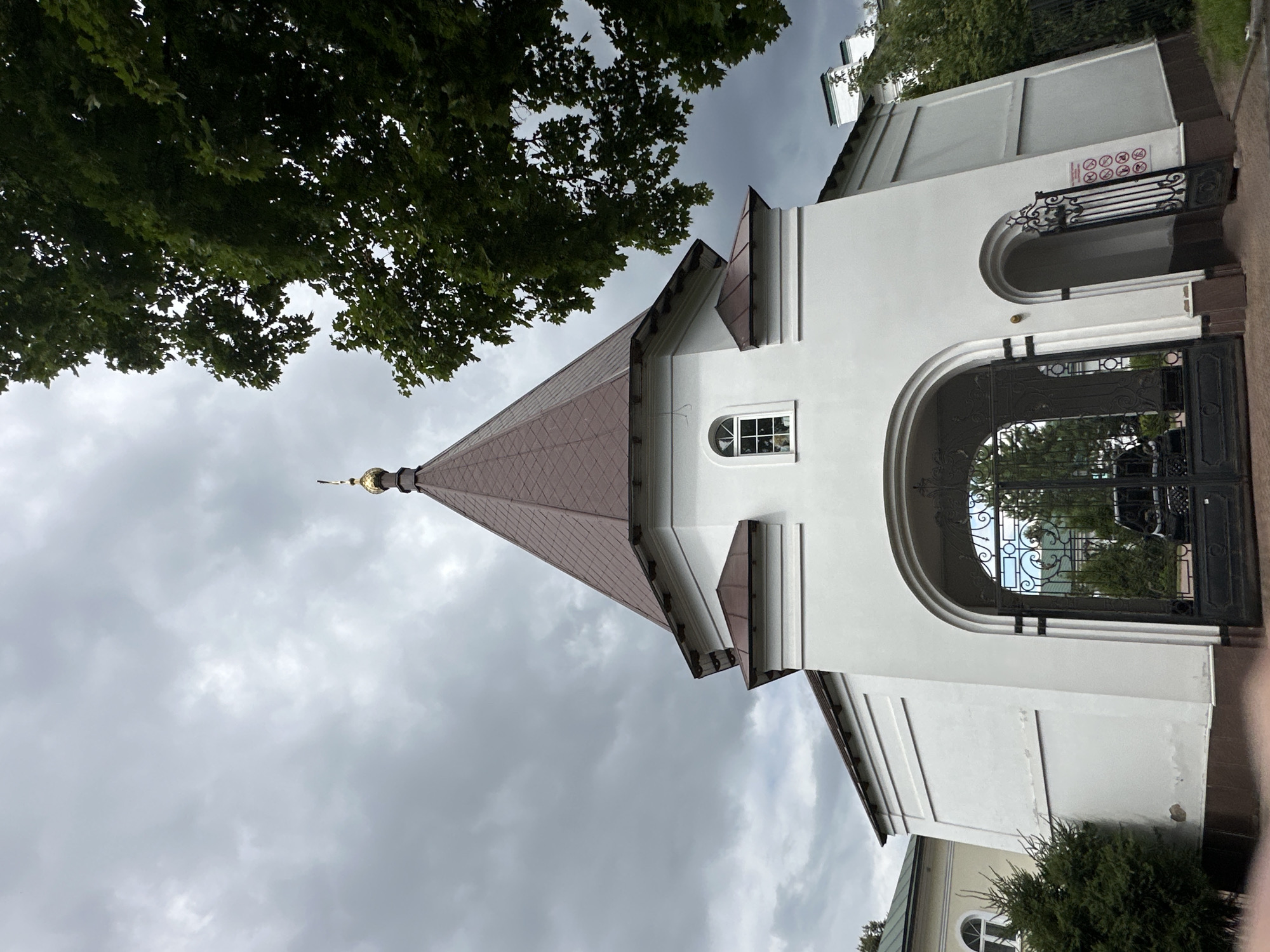 The image shows the entrance gate to a complex in Podolsk, Russia, under a cloudy sky. The gate is made of black wrought iron and has an ornate design. It leads to a courtyard where a black car is parked. To the left of the gate is a white building with a pitched brown roof, topped with a golden dome. A small arched window is visible on this building. To the right of the gate, there is a smaller arched entrance with a similar wrought iron gate. A sign with multiple circular red icons indicating prohibitions is posted on the wall next to this smaller entrance. Green trees and bushes surround the gate and buildings. In the background, more white buildings with pitched roofs and chimneys are visible, along with more trees. The overall impression is of a religious or historical complex.