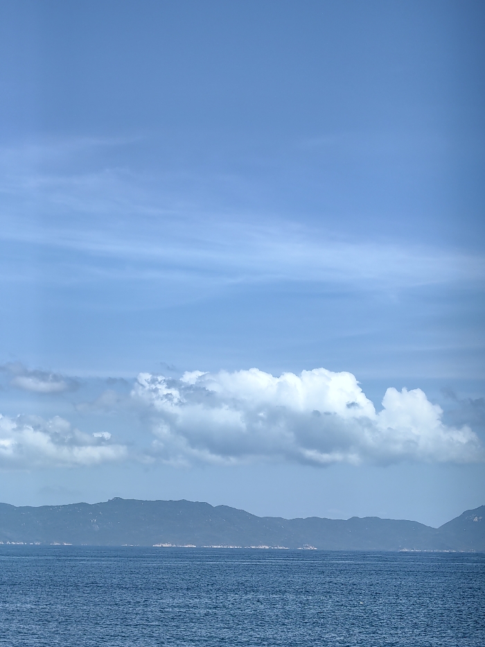 A serene seascape displays a calm, blue ocean beneath a bright, expansive sky. Wispy cirrus clouds stretch across the upper portion, transitioning to a thick band of puffy cumulus formations below. A dark, mountainous landmass sits distantly at the horizon, seemingly floating above the water. The scene conveys tranquility and vastness.