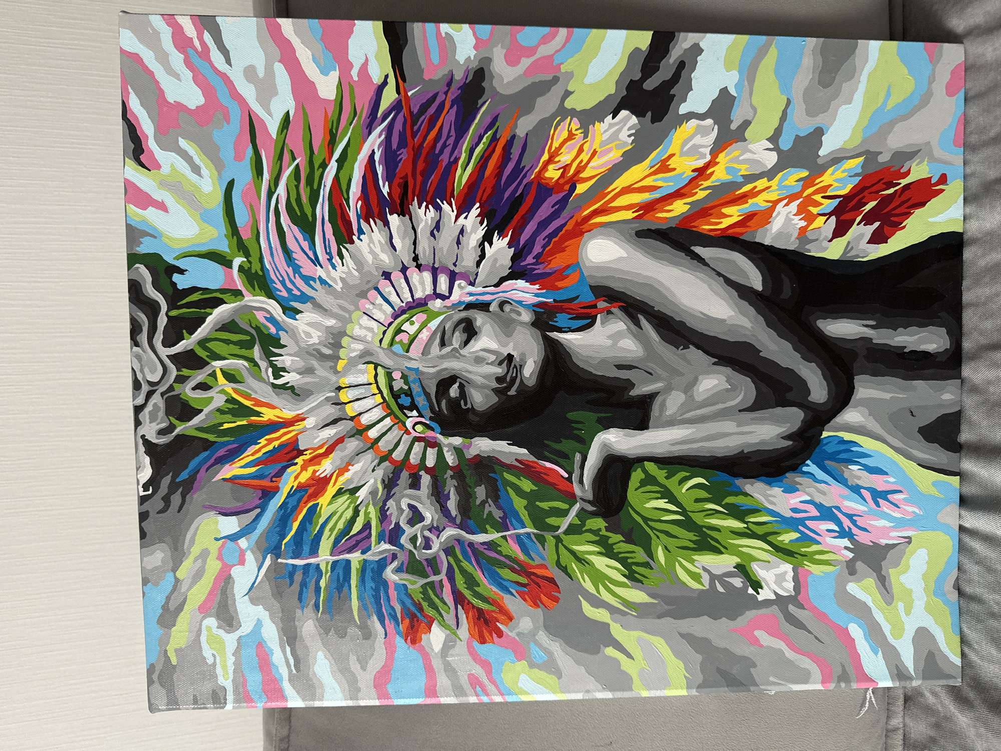 The image displays a paint-by-numbers canvas featuring a portrait of a person adorned with a large, colorful headdress reminiscent of indigenous American war bonnets. The headdress is composed of numerous feathers in vibrant shades of red, orange, yellow, green, blue, purple, and white. The central figure is rendered in grayscale, with the face in profile, eyes closed, and a cigarette held between the fingers of the left hand, with smoke trailing upwards. The background is a chaotic abstract pattern of swirling colors including gray, light blue, pink, green, and black. The canvas is positioned vertically on a gray surface, likely a couch, with a white wall visible in the upper background. The image does not contain any information about Znamya Oktyabrya, Russia.