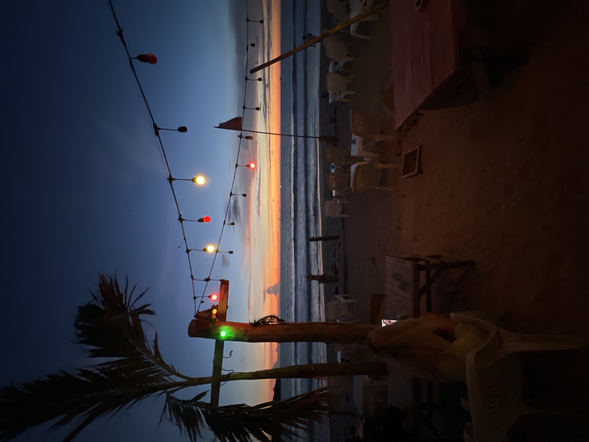 This is a vertical, eye-level, medium shot of a beach restaurant at dusk. The sky is a deep blue, transitioning to orange and pink hues at the horizon. A string of colorful lights hangs across the scene, with red, yellow, and green bulbs illuminated. A palm tree is on the left, silhouetted against the sky. In the foreground, a woman with blonde hair sits facing away from the camera, looking at her phone. Beach chairs and tables are scattered on the sand. The ocean is visible in the background with gentle waves. The scene evokes a relaxed and tropical atmosphere.