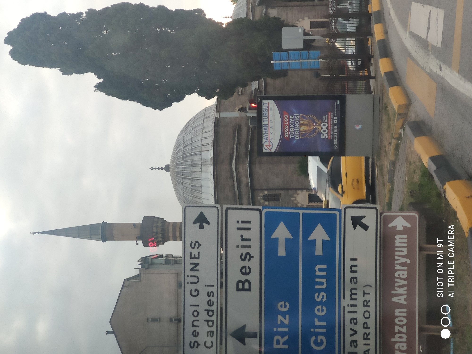 This is an eye-level, outdoor, medium shot of a street scene in Trabzon, Turkey, with a mosque visible in the background. In the foreground, there are several directional signs. The top sign, in white with black text, reads "Şenol Güneş Caddesi" with a right-pointing arrow. Below that, another white sign with black text reads "Beşirli" with an upward-pointing arrow. The next two signs are blue with white text and arrows. The first reads "Rize" with a right-pointing arrow, and the second reads "Giresun" with a right-pointing arrow. The following sign is white with black text and a black arrow pointing to the right, reading "Havalimanı (AIRPORT)". The bottom sign is brown with white text and a right-pointing arrow, reading "Trabzon Akvaryum". To the right of the signs, a billboard displays an advertisement. A yellow taxi and a white vehicle are parked on the street. The background features a mosque with a dome and a tall minaret. The sky is overcast.