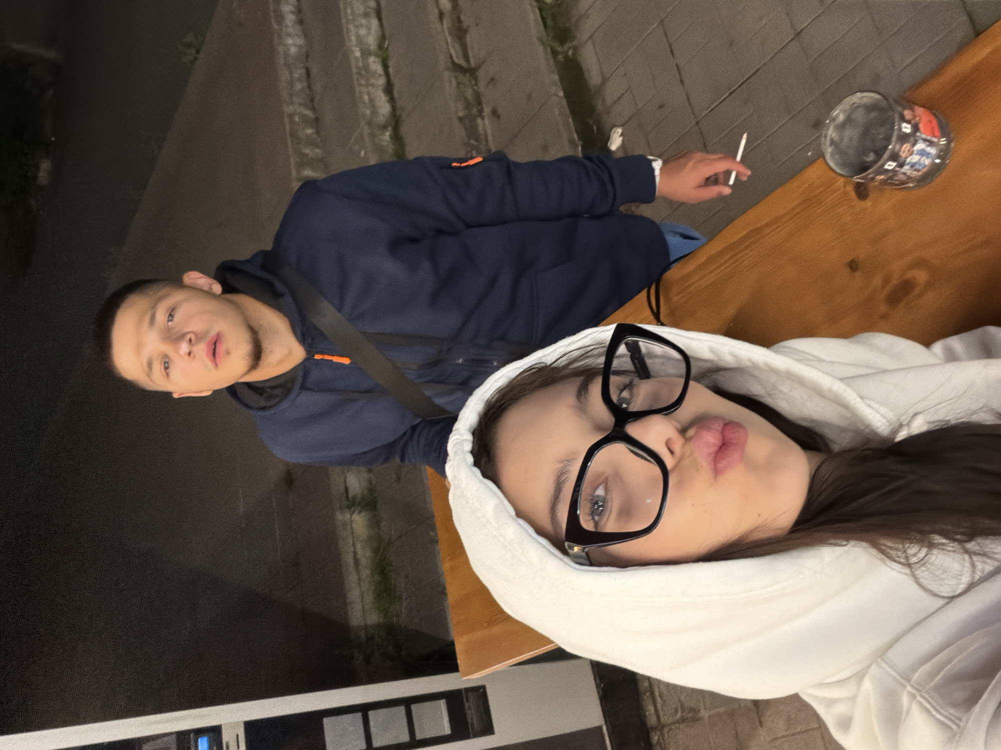 In Sofia, Bulgaria, a young woman with long dark hair, wearing a white hooded sweatshirt and large black-rimmed glasses, is taking a selfie. She is puckering her lips and leaning towards the camera, with her hooded sweatshirt pulled up around her head. To her right, a young man wearing a dark blue hooded sweatshirt stands slightly behind her. He is looking towards the camera with an open mouth. He has a dark crossbody strap over his sweatshirt. A white cigarette is visible in his right hand. On the wooden table in front of them, a cylindrical cup decorated with images of people is placed. The scene appears to be outdoors at night, with a dimly lit background showing concrete steps and a sidewalk. A tall, vertical machine or structure is partially visible on the far left of the image.