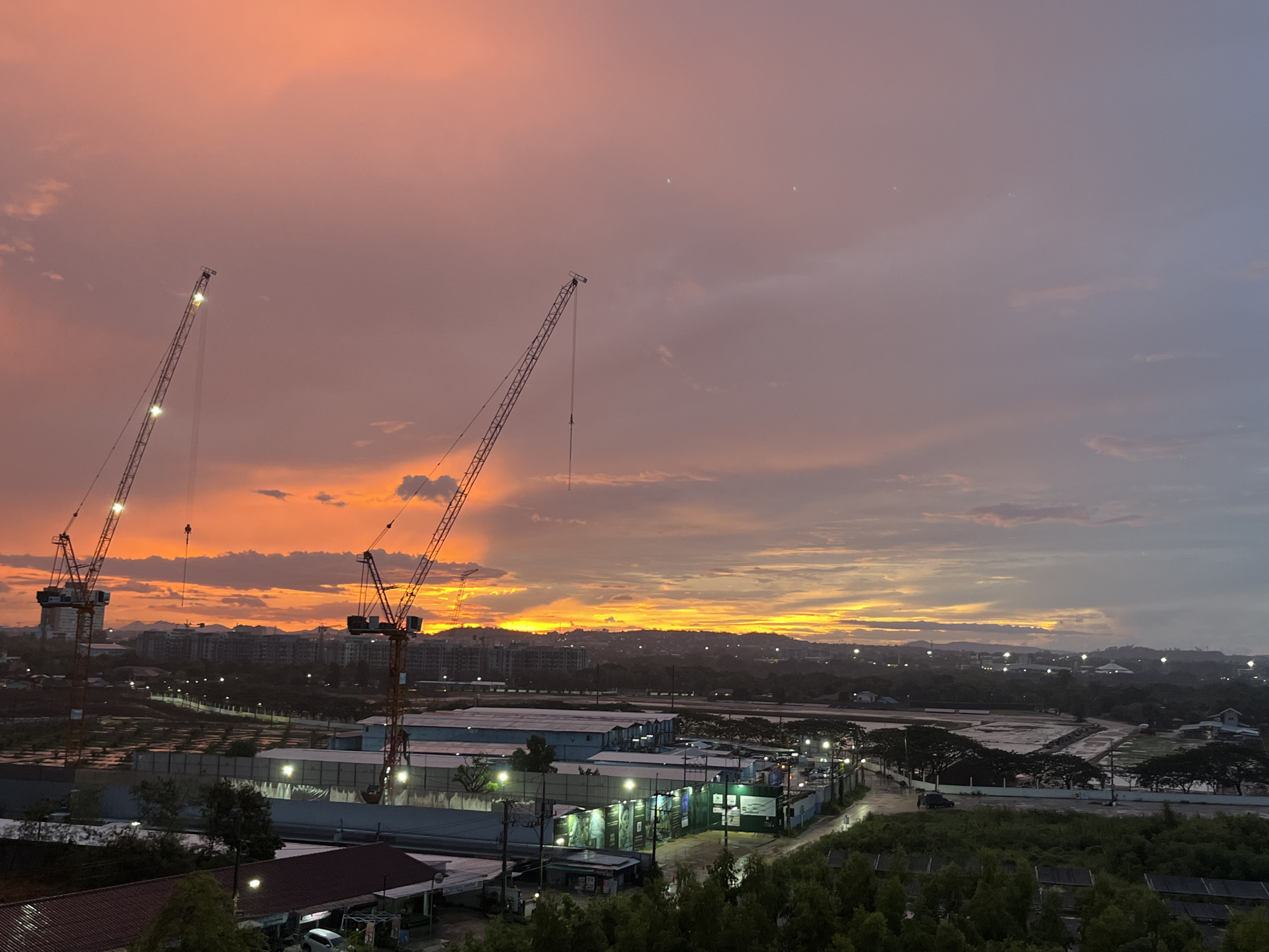 The image depicts an elevated view of a landscape, likely in Pattaya City, Thailand, given the title. The scene is set at dawn or dusk, as indicated by the vibrant orange and pink hues of the sky. Several construction cranes are visible in the foreground, suggesting ongoing development in the area. Below the cranes, there is a cluster of industrial or commercial buildings illuminated by streetlights. In the background, a cityscape fades into the horizon with a range of buildings. The environment appears to be a mix of urban and natural elements, with trees and greenery interspersed among the buildings. No people are directly observable.