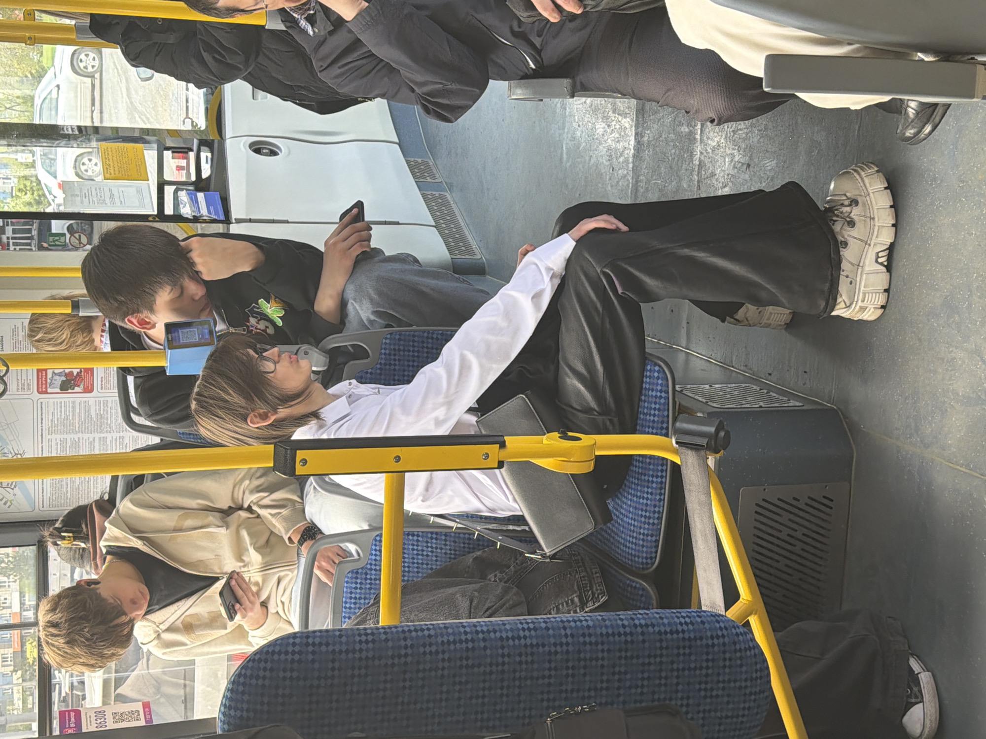 The image depicts the interior of a public bus, likely a city bus, possibly located in Krasnoyarsk, Russia, given the Cyrillic text visible on a poster. Several people are seated, some using their phones. The seating has blue patterned upholstery, and the bus features yellow handrails. Passengers are dressed in casual wear, including jackets, sweatshirts, and pants. One individual wears a white shirt. An exterior view through the bus windows reveals buildings, parked cars, and greenery, suggesting an urban environment. The scene is captured from an elevated perspective within the bus.