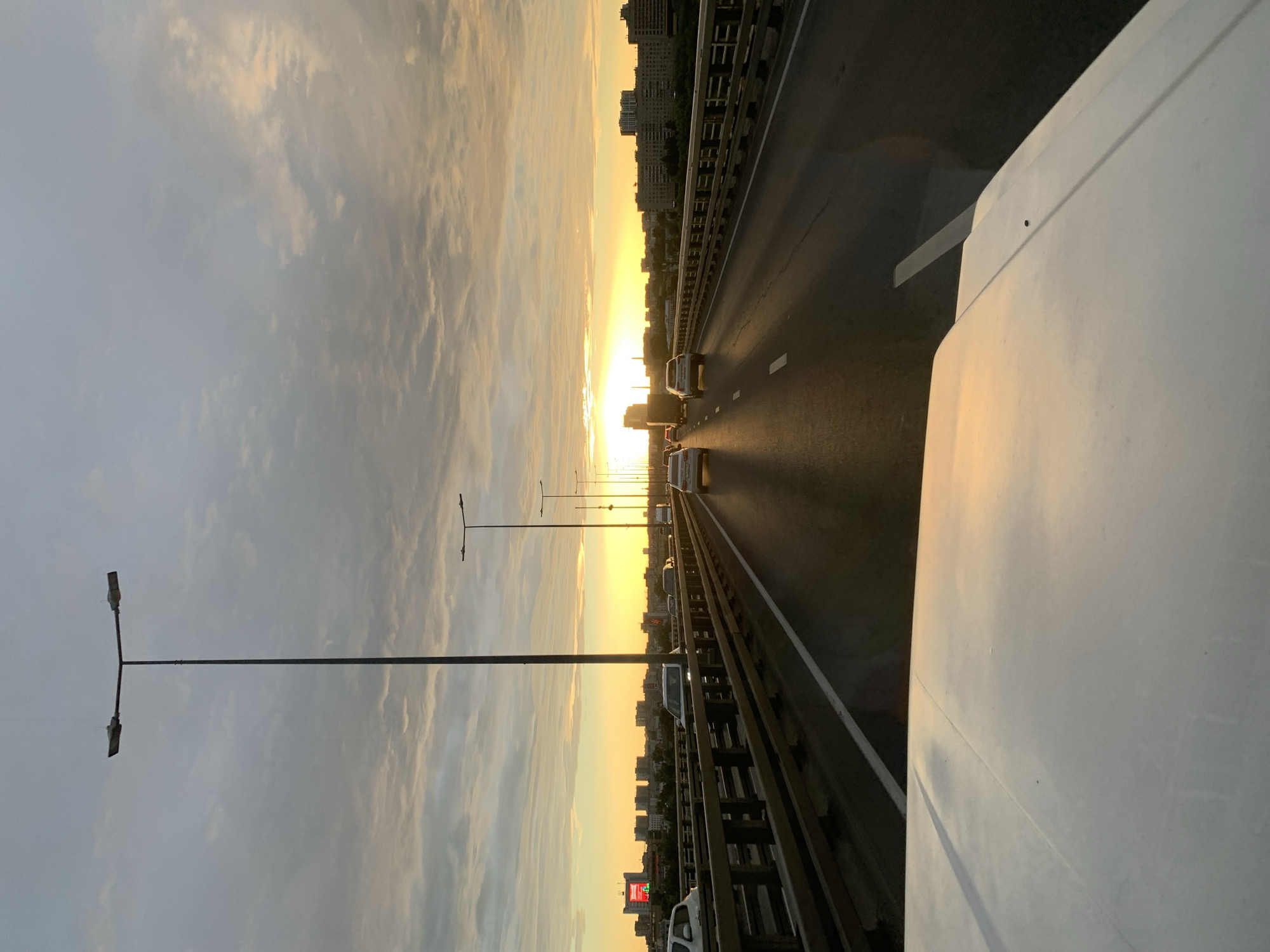 A view from the front of a white vehicle traveling over a bridge during sunset. The sky is filled with clouds, with the sun setting on the horizon and casting a warm, golden glow over the scene. A city skyline is visible in the distance, with buildings of various heights. Multiple cars are driving on the bridge, with the vehicle from which the photo is taken being the closest to the foreground. Tall streetlights line the sides of the bridge. The railing of the bridge is made of wood and metal. The scene suggests a commute or travel through the city during the evening.