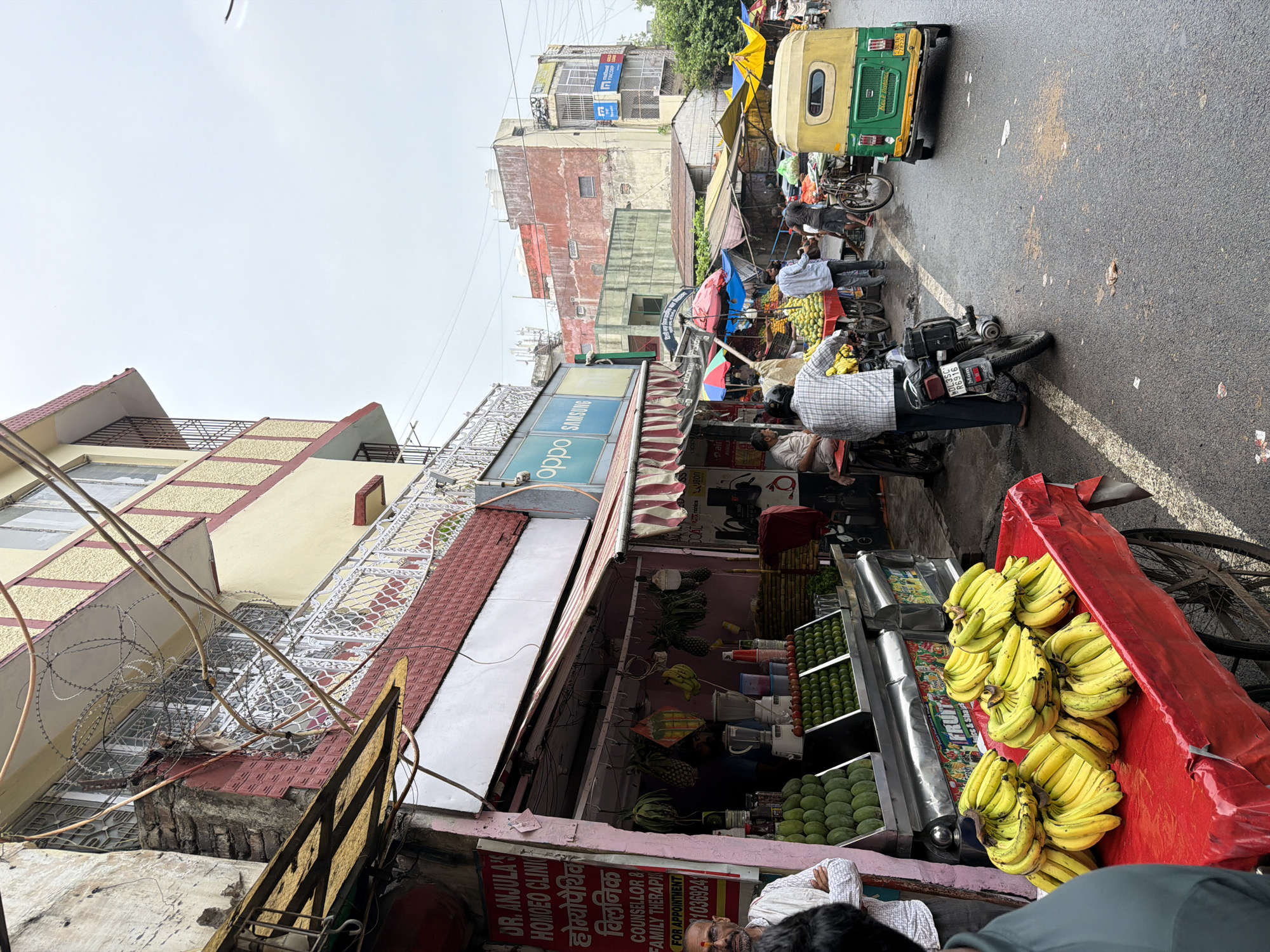 The image depicts a street scene in Delhi, India, likely a market area, characterized by a bustling atmosphere with vendors and shoppers. A street vendor is seen displaying a large quantity of ripe bananas on a red cart covered with plastic. To the left, a homeopathic clinic sign is visible, with "DR. ANJULA'S HOMOEOPATHIC CLINIC" written in English and Hindi. The clinic also advertises "Counsellor & Family Therapies." A man wearing glasses and a light-colored shirt stands near the clinic's entrance, appearing to be a customer or bystander. Behind the bananas, stalls are set up selling various fruits, including mangoes and what appear to be limes, arranged in crates. Several people are present, some tending to the stalls, while others are walking or standing. A yellow and green auto-rickshaw is parked on the right side of the street, along with a motorcycle. The street is paved and appears wet, suggesting recent rain. The buildings in the background are multi-story structures, some showing signs of age and weathering. The overall scene captures the vibrant and active street life typical of a market in Delhi.