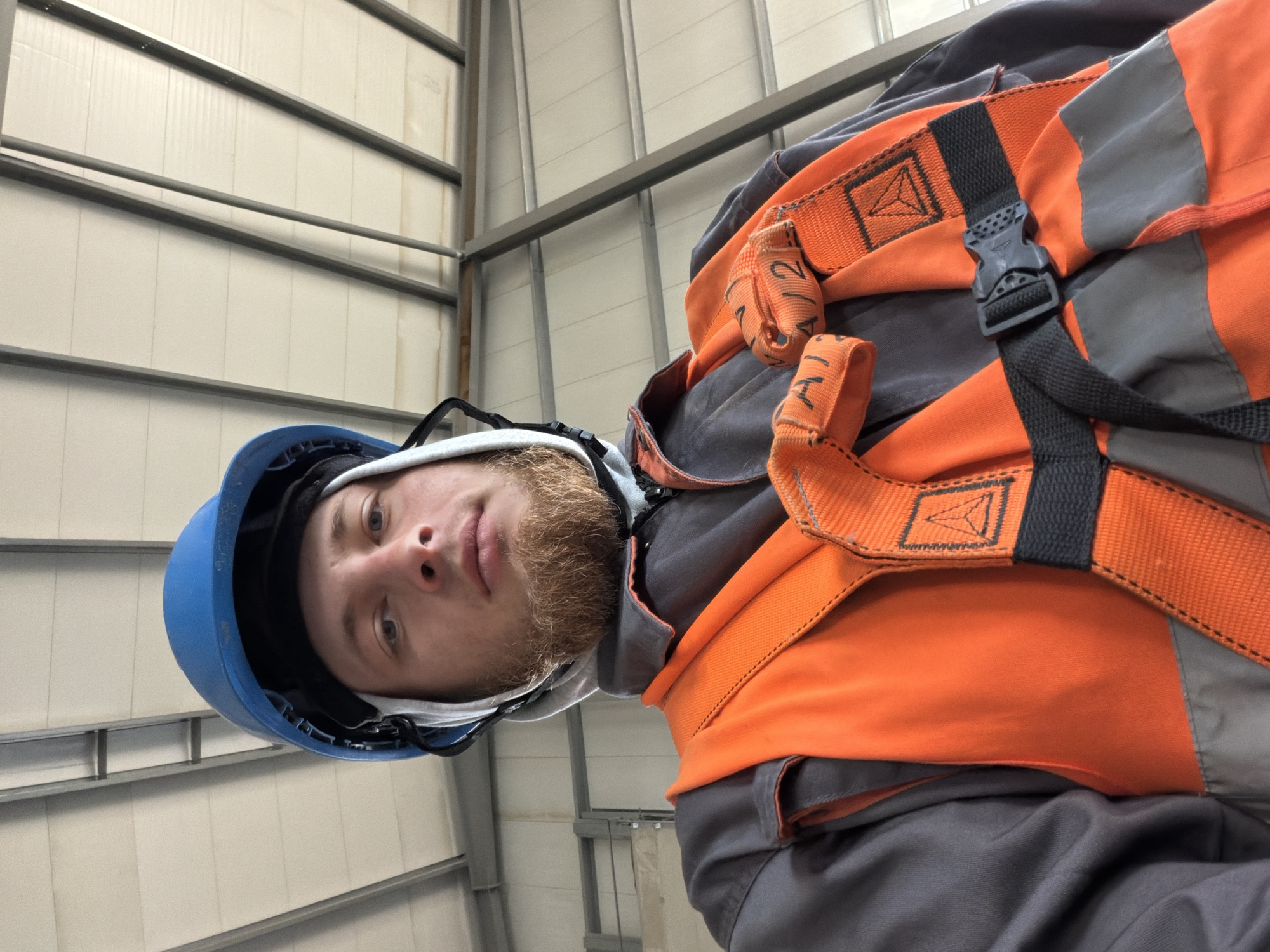 A man with a beard wearing a blue hard hat and an orange safety vest is looking up at the camera from inside a building with a visible ceiling structure. The man is wearing a grey work jacket underneath the vest. The background consists of the interior of a building with a metal frame and a white ceiling. The location is Jodłownik, Poland.