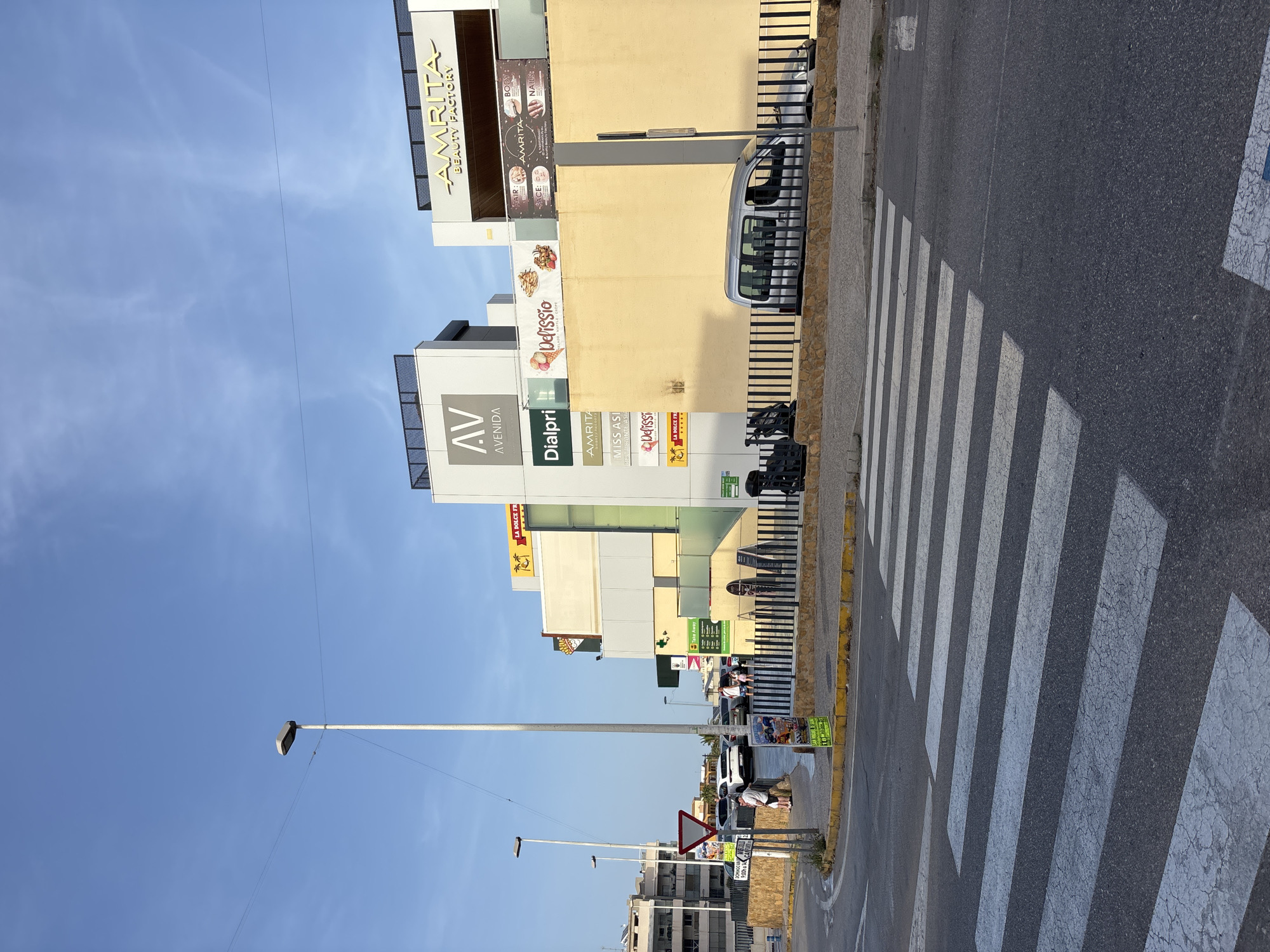 The image captures a street scene in Urbanización Villa Martín, Spain, on a clear day with a blue sky and scattered clouds. In the foreground, a pedestrian crosswalk is clearly marked on the asphalt road. To the left, a white SUV is parked near a roundabout with a yield sign. Beyond the roundabout, several apartment buildings are visible.
Dominating the midground and background is a multi-story commercial building complex with various signs and storefronts. Prominent signage includes "AV AVENIDA" on a light grey facade, and "-AMRITA BEAUTY FACTORY-" in gold lettering on a white structure. Below these, other signs indicate businesses such as "Dialpri," "Delfissio" (offering "HELADO WAFFLE CREPE"), and "MISS ASI RESTAURANTE ASI." A green sign for "Take Away" is also visible.
A black metal fence separates the sidewalk from the street in front of the commercial buildings. A silver minivan is parked behind the fence, adjacent to the building. Several individuals are present, including two people standing near the parked SUV, and three men walking near the commercial establishments. The overall impression is of a sunny day in a commercial area.