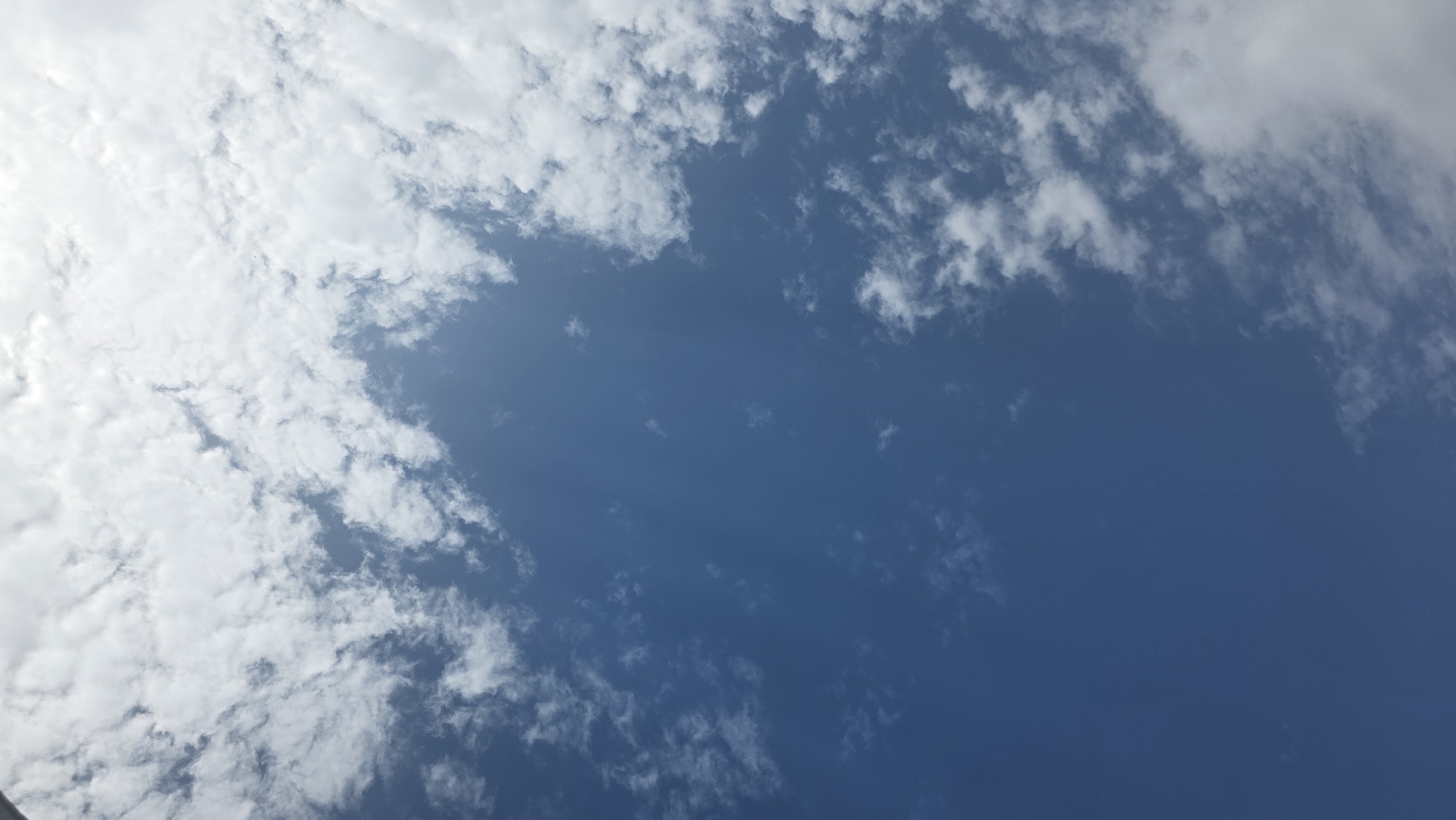 The image shows a clear blue sky with scattered white clouds. The clouds are wispy and varied in shape and size, with some appearing thicker and more substantial than others. The lighting suggests it is daytime, with the sun likely overhead or slightly to the side, casting a bright glow on the clouds. There are no discernible buildings, people, or objects within the frame, indicating an unobstructed view of the sky. The absence of specific landmarks or environmental features makes it impossible to definitively place this image within Tokmok City, Kyrgyzstan, or any other specific location.