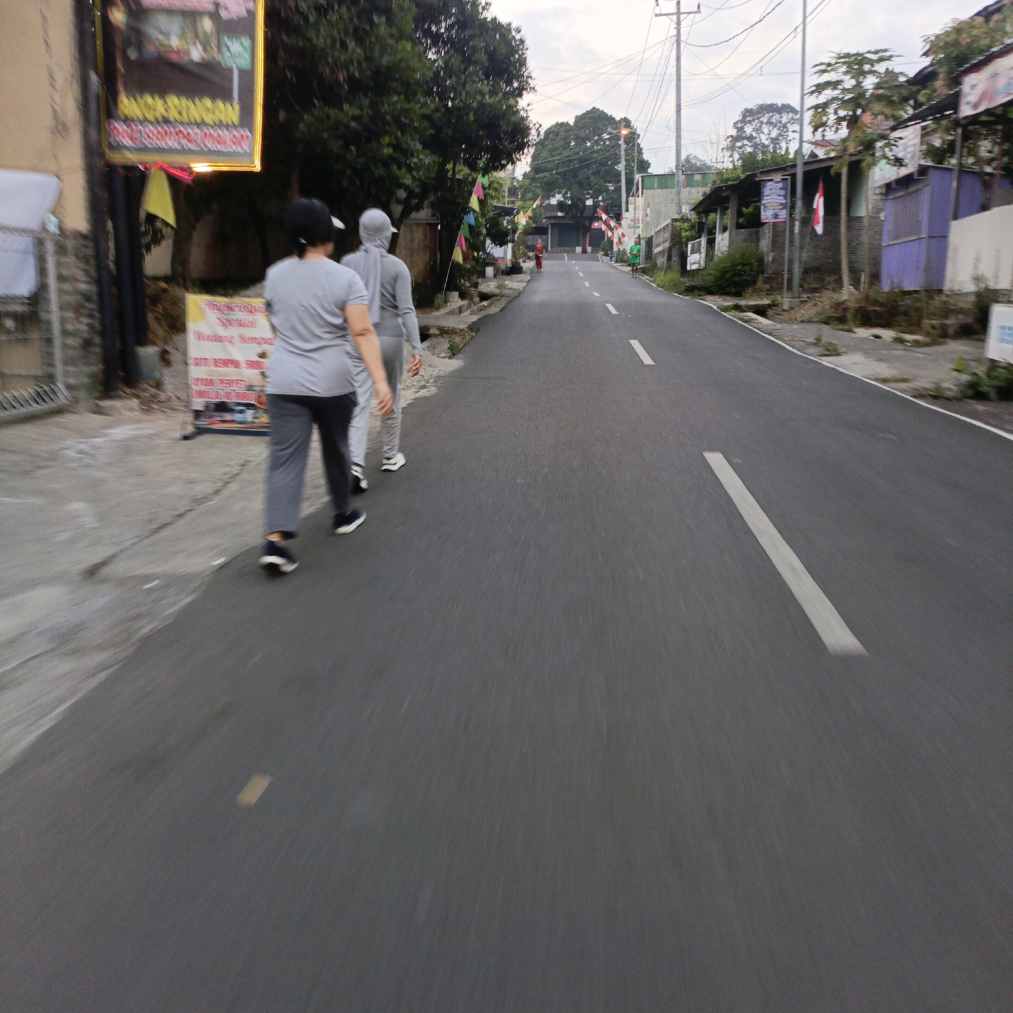 The image shows a street scene in Salatiga, Indonesia, with a focus on a road and several people walking. In the foreground, two individuals are walking away from the camera on the left side of the road. The person closer to the camera is wearing a grey t-shirt and dark pants with black sneakers. The second person is wearing a grey hooded outfit and a headscarf, also with dark pants and white sneakers. To the left of the road, a yellow sign with red and black text is visible, mounted on a pole with a green banner above it. Further down the road, two more individuals are walking in the same direction as the foreground figures. One is wearing a red outfit, and the other is in a green outfit. The road is paved with white dashed lines marking the lanes. Buildings and trees line the sides of the road, with some appearing to be shops or houses. Several Indonesian flags are also visible, some attached to poles and others displayed on buildings. The sky is overcast, suggesting it might be early morning or late afternoon. The scene appears to be a public street in a residential or commercial area of Salatiga.