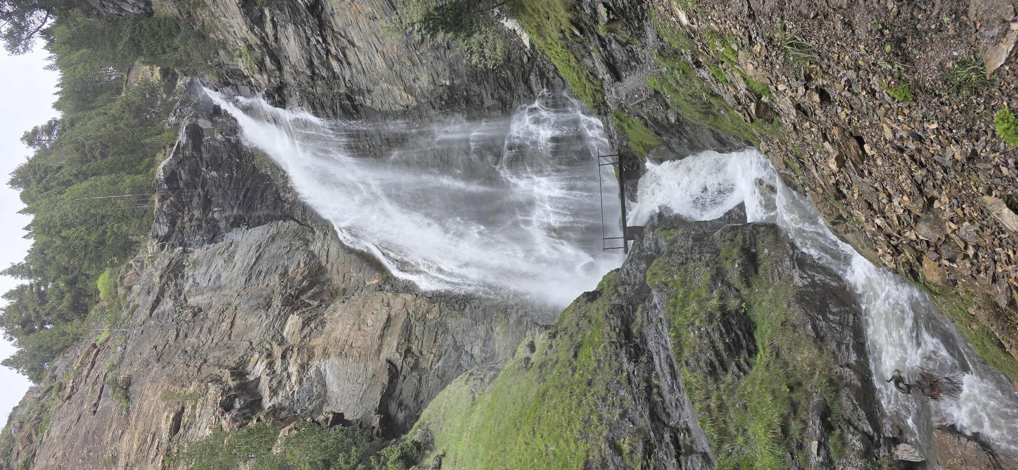 The image shows the Salto de Paderna waterfall in Benasque, Spain. Water cascades down a rocky cliff face, surrounded by green vegetation. A metal structure is visible mid-cascade. The surrounding environment consists of rocks, grass, and trees.