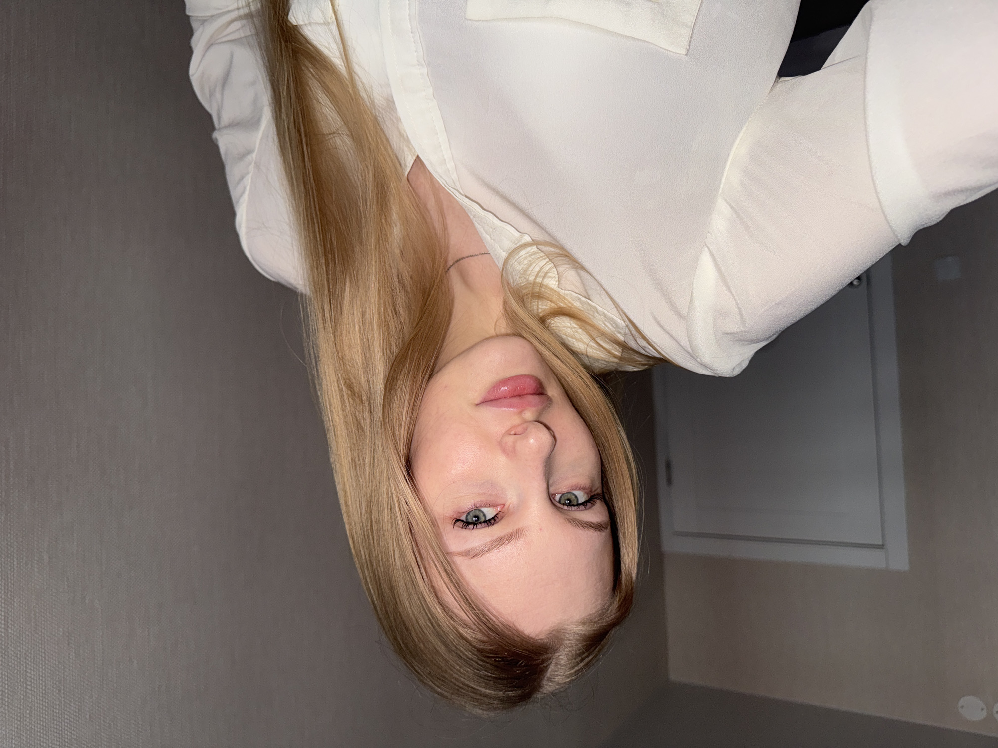 A young woman with long, straight blonde hair is wearing a white, long-sleeved blouse. The image is taken indoors, likely in a room with gray walls and a door visible in the background. The subject is facing the camera, though the image has been rotated 180 degrees. No city or specific location is discernible.