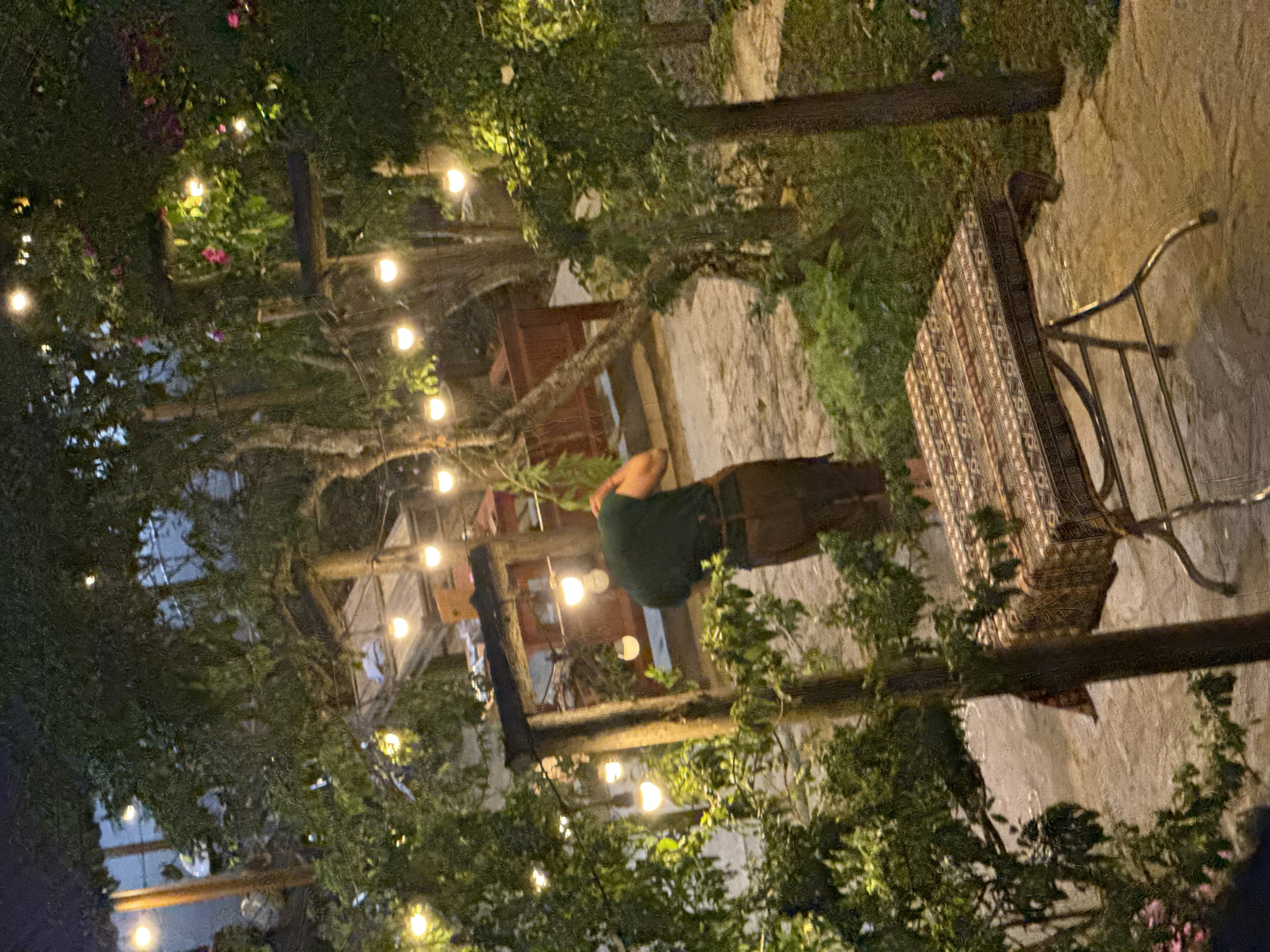 The photograph shows an outdoor scene in Bắc Hà, Vietnam, at night. String lights illuminate a patio area with trees and foliage. A person wearing a green shirt and brown pants stands near a table with a patterned tablecloth. The ground is stone or concrete. The area appears to be a garden or patio setting.