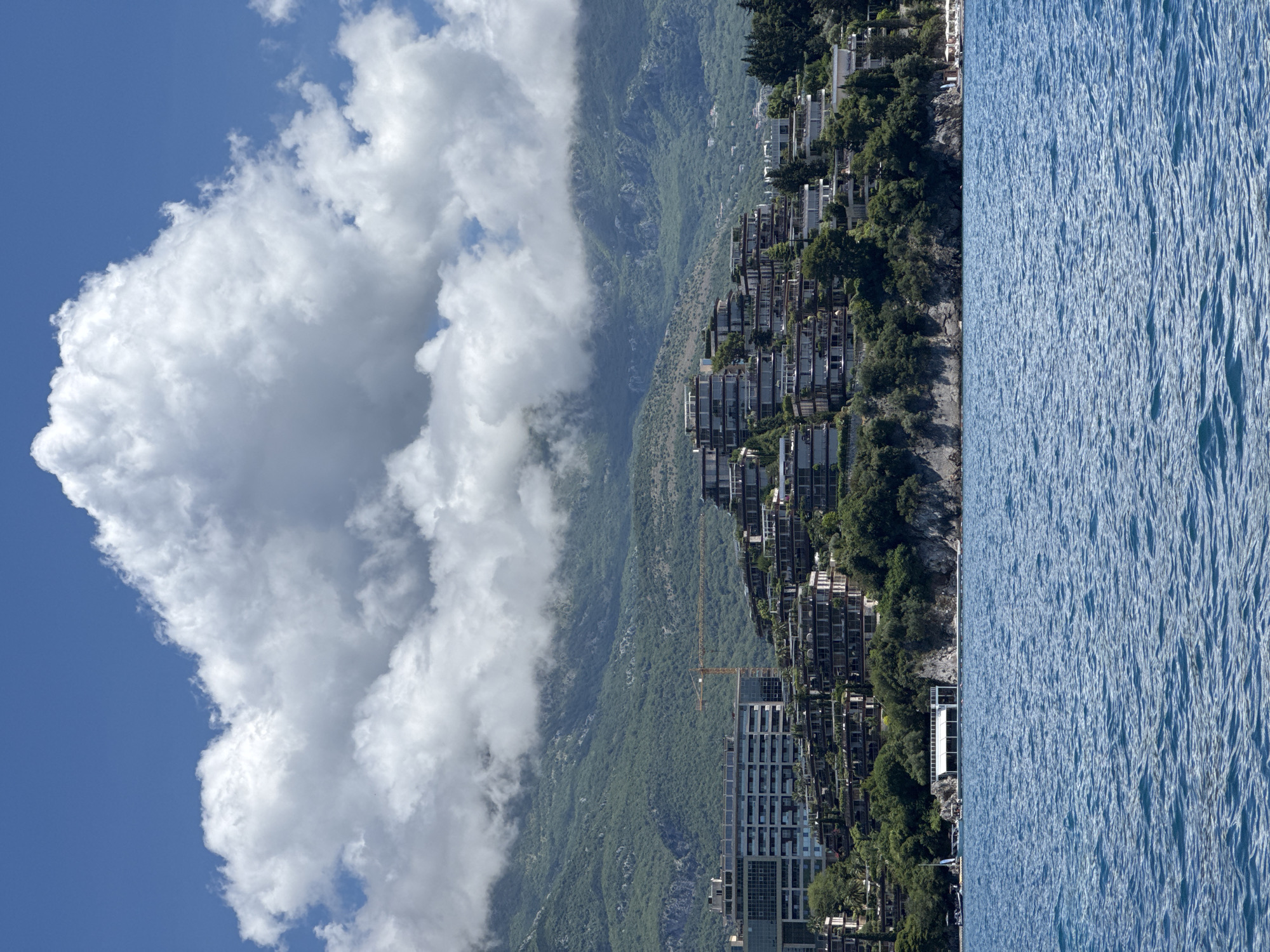 The image, when correctly oriented, presents a panoramic view of a bustling coastal landscape. In the foreground, a vast expanse of blue water, likely the Adriatic Sea, stretches across the bottom of the frame, its surface textured with countless small, shimmering ripples.
Above the water line, a verdant coastline rises, heavily developed with numerous modern, multi-story buildings. These structures, appearing to be residential complexes or hotels, are built into and along the green, tree-covered slopes. Lush green foliage, including trees and shrubs, dots the terrain between the buildings. Evidence of ongoing construction is visible, with scaffolding on some buildings and a prominent yellow crane standing tall in the mid-ground, indicating active development.
Behind this developed strip, a majestic mountain range dominates the background. Its slopes are densely covered in rich green vegetation, with rocky outcrops visible in places. Large, white, fluffy cumulus clouds partially obscure the higher peaks of the mountains, adding depth to the scene. The sky above is a clear, bright blue, populated by more white clouds.
The scene depicts a sunny day, likely late morning or early afternoon, with excellent visibility. While no specific human activity is shown, the extensive development suggests a vibrant and populated coastal area, typical of a resort town. The overall impression is one of a scenic and dynamic coastal environment. There is no visible text in the image.