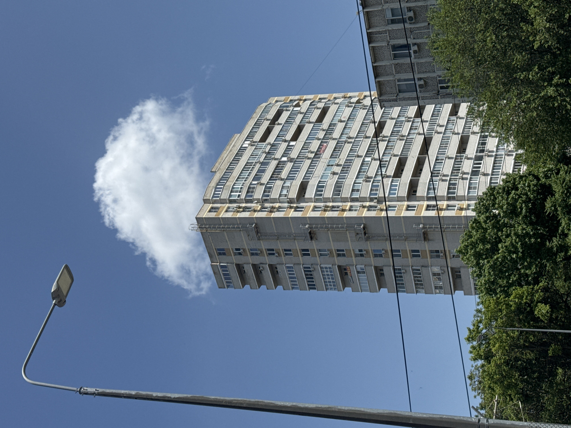 The image shows a tall, Soviet-era apartment building in Krasnodar, Russia, viewed from a low angle. The building is gray and boxy, with rows of windows and air conditioning units. A puffy white cloud appears next to the building, seemingly level with its top. To the right of the building is a large green tree, and in the foreground is a gray streetlight and power lines against a clear blue sky.