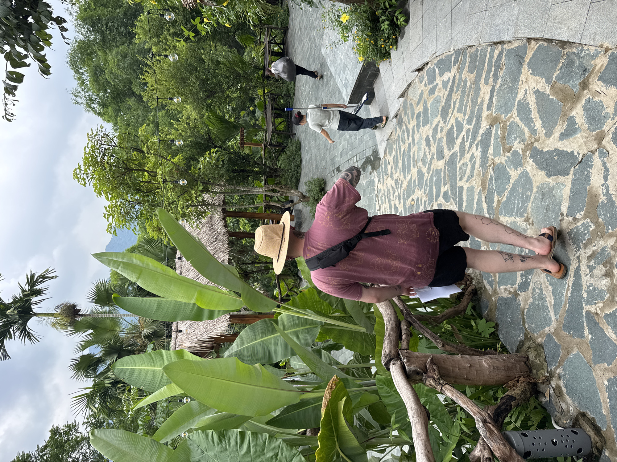 The image shows an outdoor scene in Pù Luông Commune, Vietnam. A person wearing a straw hat, a maroon t-shirt, and black shorts is walking away from the camera on a stone pathway. They have tattoos on their legs. To the left of the pathway is a wooden railing and lush green foliage, including large leaves. In the background, there are thatched-roof structures and more greenery, suggesting a resort or natural park setting. Further down the pathway, two other individuals are visible. One is a young person holding a broom and a dustpan, walking towards the camera. Another person is walking away from the camera in the distance, carrying a large grey bag. The sky is overcast.