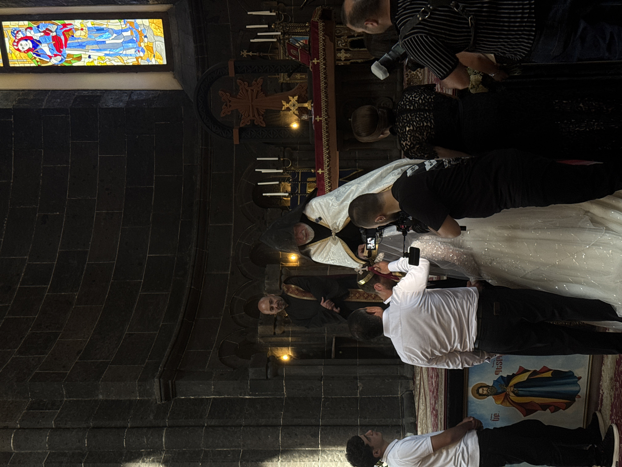 In the city of Gyumri, Armenia, inside an Armenian Apostolic church, a wedding ceremony is taking place. A priest, dressed in a white and gold robe and a conical hat, officiates the ceremony. He is standing in front of an altar adorned with religious symbols, candles, and a bible. A bride in a white, sparkling wedding dress and a groom in a white shirt and dark pants are the central figures, with the priest placing a crown on the groom's head. Several guests are present, including men in formal wear and women in elegant dresses. Some guests are holding cameras and recording the event. The church interior is characterized by dark stone walls, arched ceilings, and stained-glass windows depicting religious scenes. The overall atmosphere suggests a solemn and significant occasion.