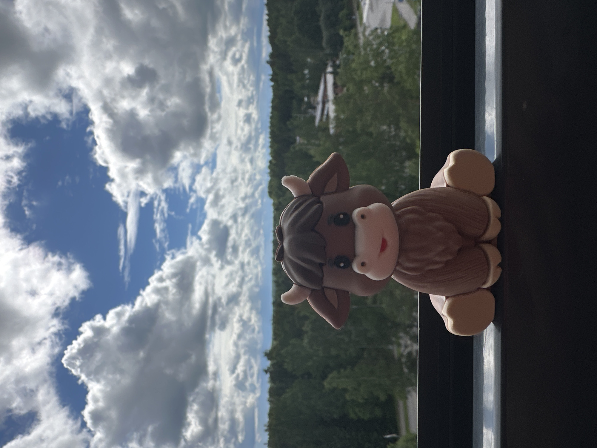 This media file shows a close-up of a small, brown, plush toy cow placed on a dark surface, likely a windowsill or balcony railing. The cow has a light brown muzzle with pink cheeks and a red mouth, black eyes, and light brown ears. Its body is a darker brown, textured to resemble fur. The cow is positioned facing to the right, with its body resting on the surface and its head tilted slightly upwards.
In the background, a bright blue sky with fluffy white clouds is visible, suggesting daylight. Beyond the sky, a dense line of green trees forms a horizon, hinting at an outdoor or semi-outdoor setting with natural surroundings. Faint structures, possibly houses or buildings, can be seen through the trees. The overall lighting suggests it's daytime, and the clear sky with clouds indicates fair weather. The image is oriented vertically.