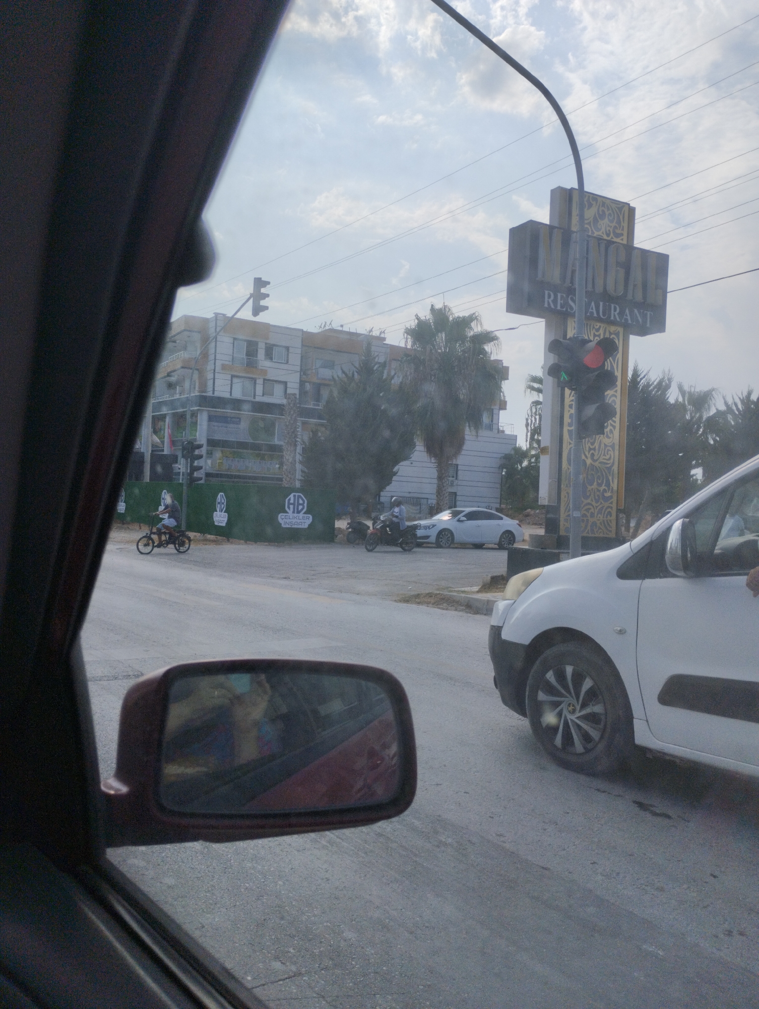 In Mezitli, Turkey, a shot taken from inside a vehicle shows a street scene. A traffic light with a red and green signal illuminated is visible. A sign for "MANGAL RESTAURANT" is mounted on a pole. In the background, there are several buildings and palm trees. In the foreground, a white van is partially visible, as is a side mirror reflecting a person inside the vehicle. A man on a bicycle rides past a green barrier. Two motorcycles and a white car are also present on the street. The sky is cloudy.