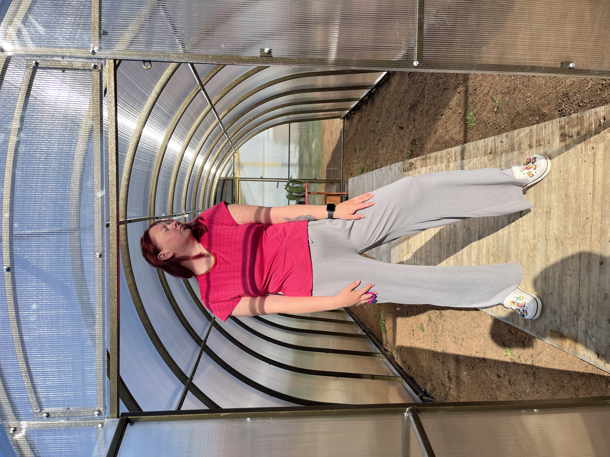 A woman lies inside a greenhouse in what is presumed to be Chaikovsky, Russia. She is wearing a pink short-sleeved shirt and gray pants, and white shoes with cartoon decorations. She is lying on her back. The greenhouse has a curved metal frame and translucent plastic walls and roof. The ground inside the greenhouse is a mixture of dirt and wooden planks. A small patch of plants grows on the ground.
