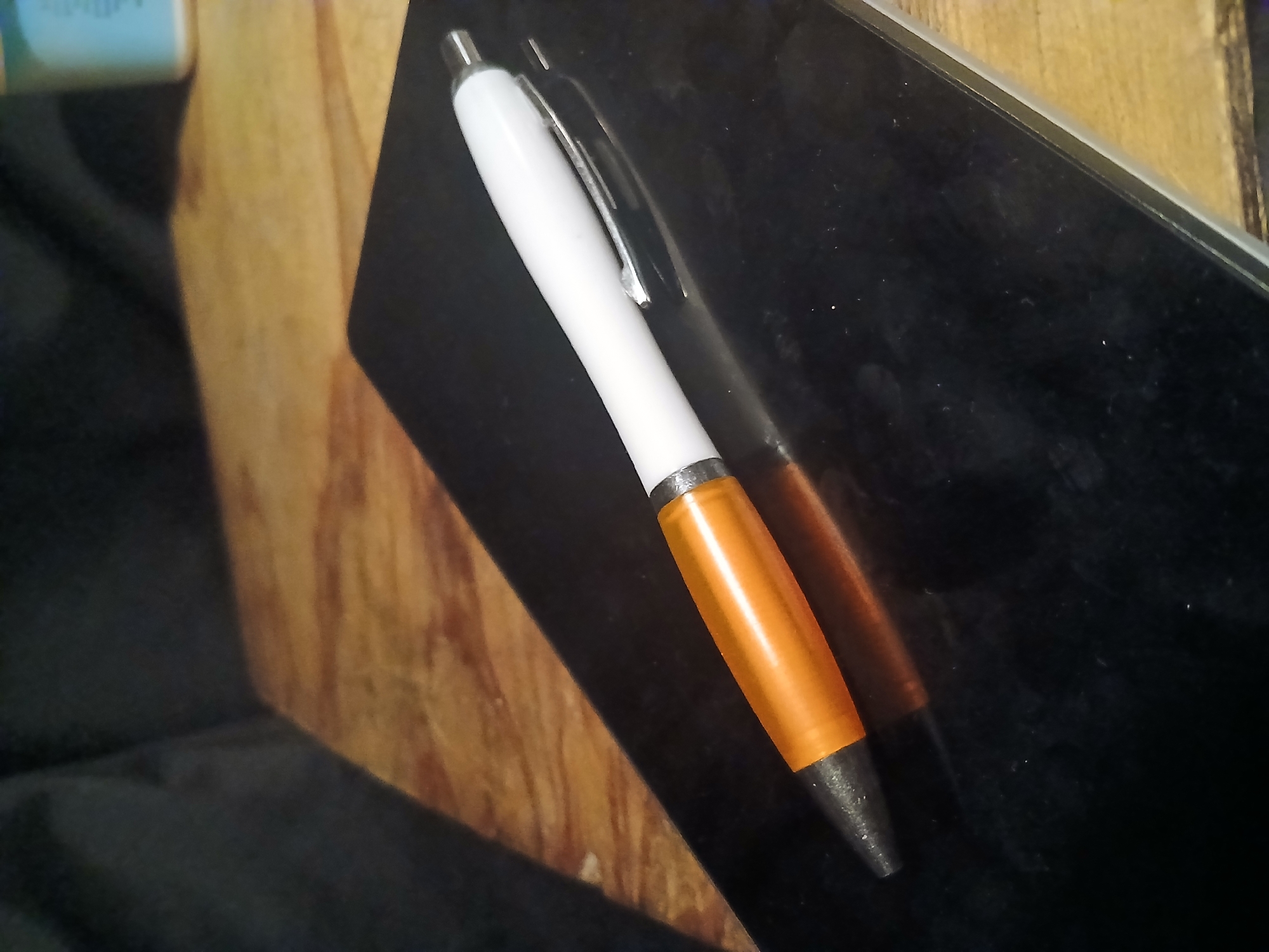 A white and orange pen rests on a black surface, possibly a tablet or e-reader. The pen is positioned diagonally, with its tip pointing towards the bottom left of the frame. The orange section of the pen appears to be a translucent grip. The black surface has a reflective quality, showing a faint reflection of the pen. The background consists of a wooden surface and a dark fabric. There is no indication of the city of Douai, France, or any specific location within it.