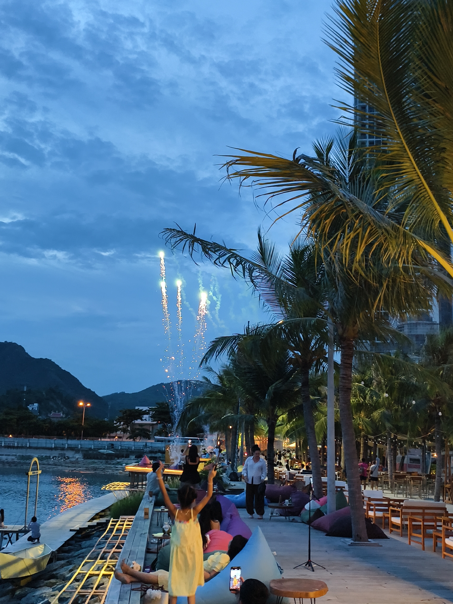 In Nha Trang Ward, Vietnam, a twilight scene unfolds by the sea, illuminated by the soft glow of distant lights reflecting on the water. Fireworks streak across the dusky sky, their ephemeral trails adding a touch of celebration to the atmosphere. Palm trees, their fronds silhouetted against the deepening blue, line the waterfront, framing a pathway dotted with people. Some individuals are seated on colorful beanbag chairs, while others stand or converse. A young girl in a yellow dress faces away from the camera, her arms raised in apparent delight at the fireworks. To her right, a man in a white shirt and dark pants walks along the path, while other patrons are visible in the background, seated at tables or strolling. The scene suggests a relaxed evening gathering, perhaps a celebration or a social event, with the natural beauty of the coast and the spectacle of fireworks creating a memorable backdrop.