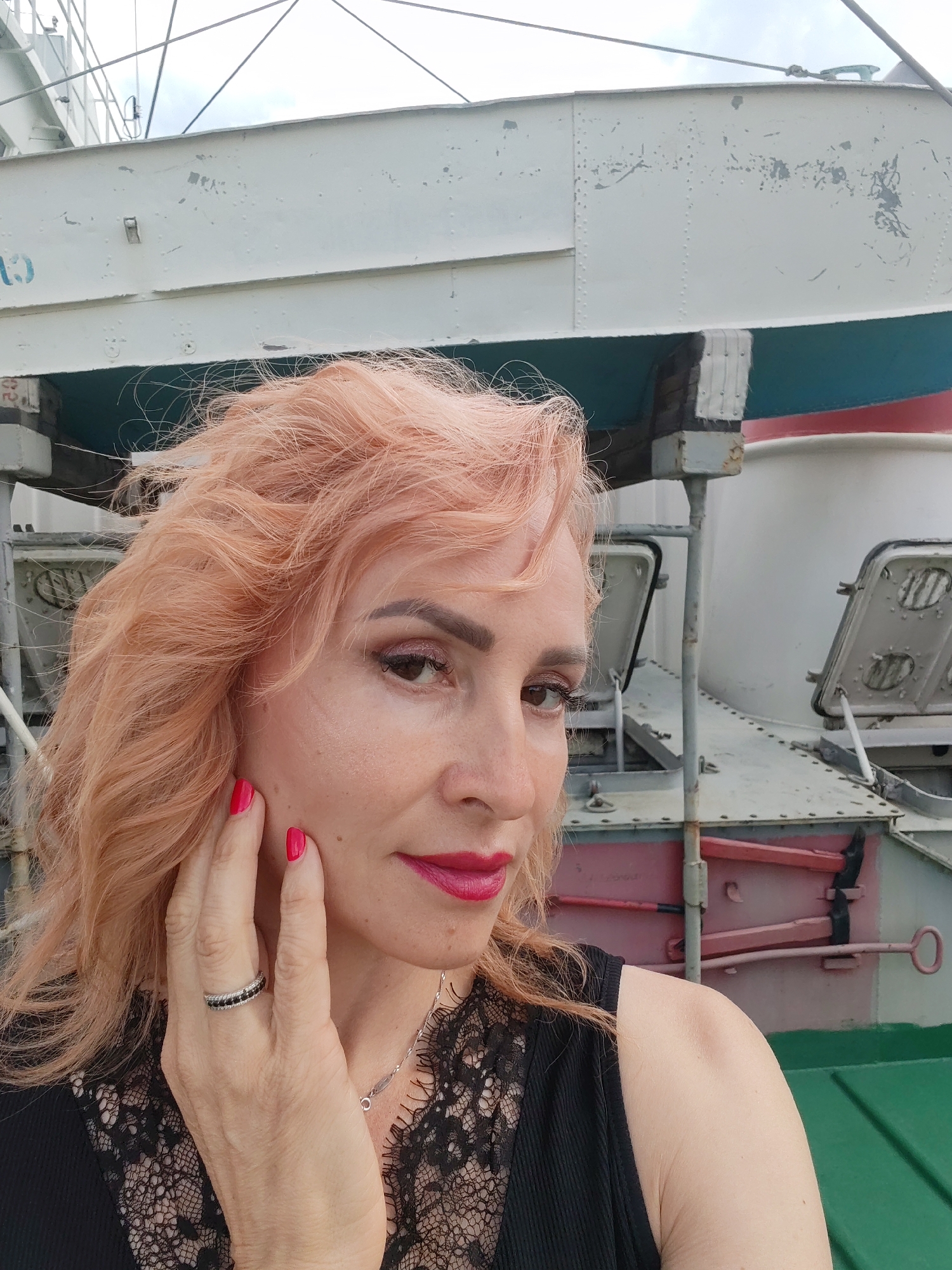 A medium shot shows a woman with peach-colored, wavy hair and bright red lipstick. She is wearing a black top with lace detailing and a silver ring with black stones on her left hand. She is touching her cheek with her left hand. The background is a light-colored metal structure with blue and red accents. The woman is positioned on the right side of the frame, looking towards the viewer. The environment appears to be outdoors, possibly a dock or industrial area. The lighting is natural and overcast. There is no information about the city of Vasil'ki, Russia, present in the image.